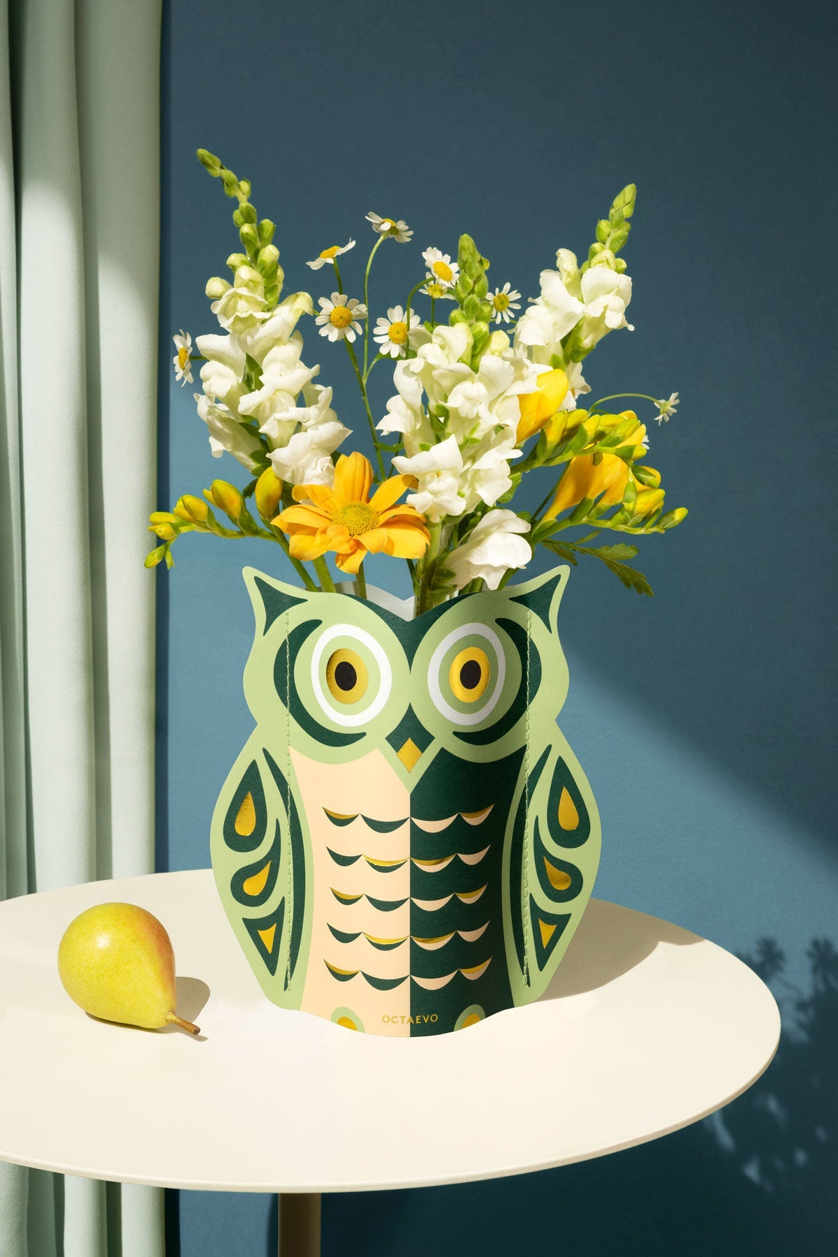 OCTAEVO Night Owl Mini Paper Vase with yellow and white blooms on a round table, set against a blue wall.