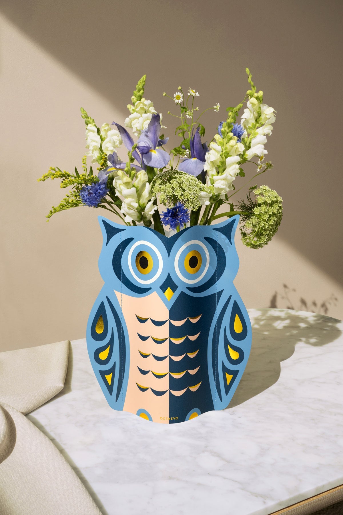 The Octaevo Night Owl Paper Vase holds assorted purple, white, and green flowers on a marble table.
