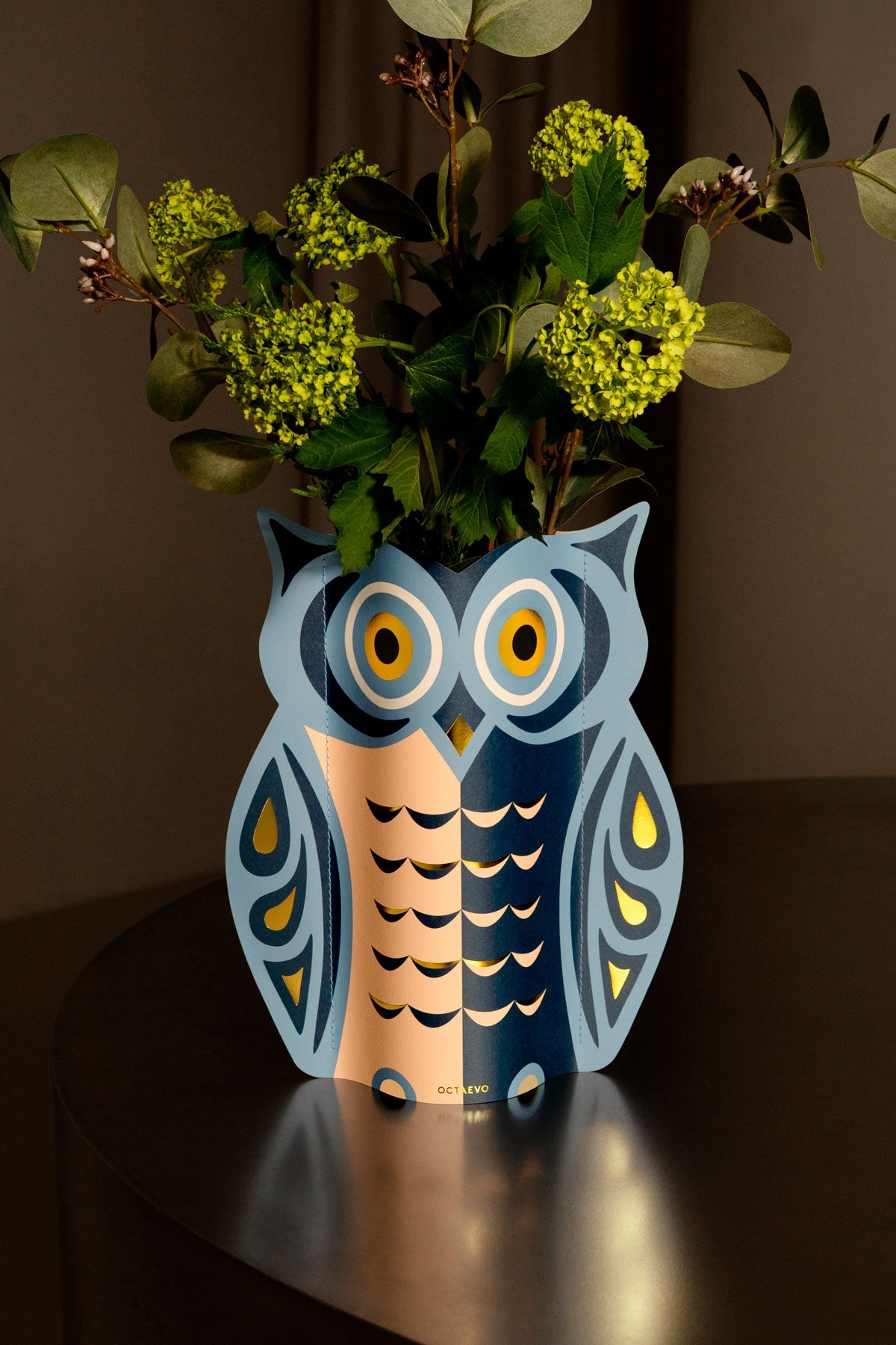 Le vase en papier Night Owl OCTAEVO accueille des fleurs vertes sur une table réfléchissante, devant un fond brun.