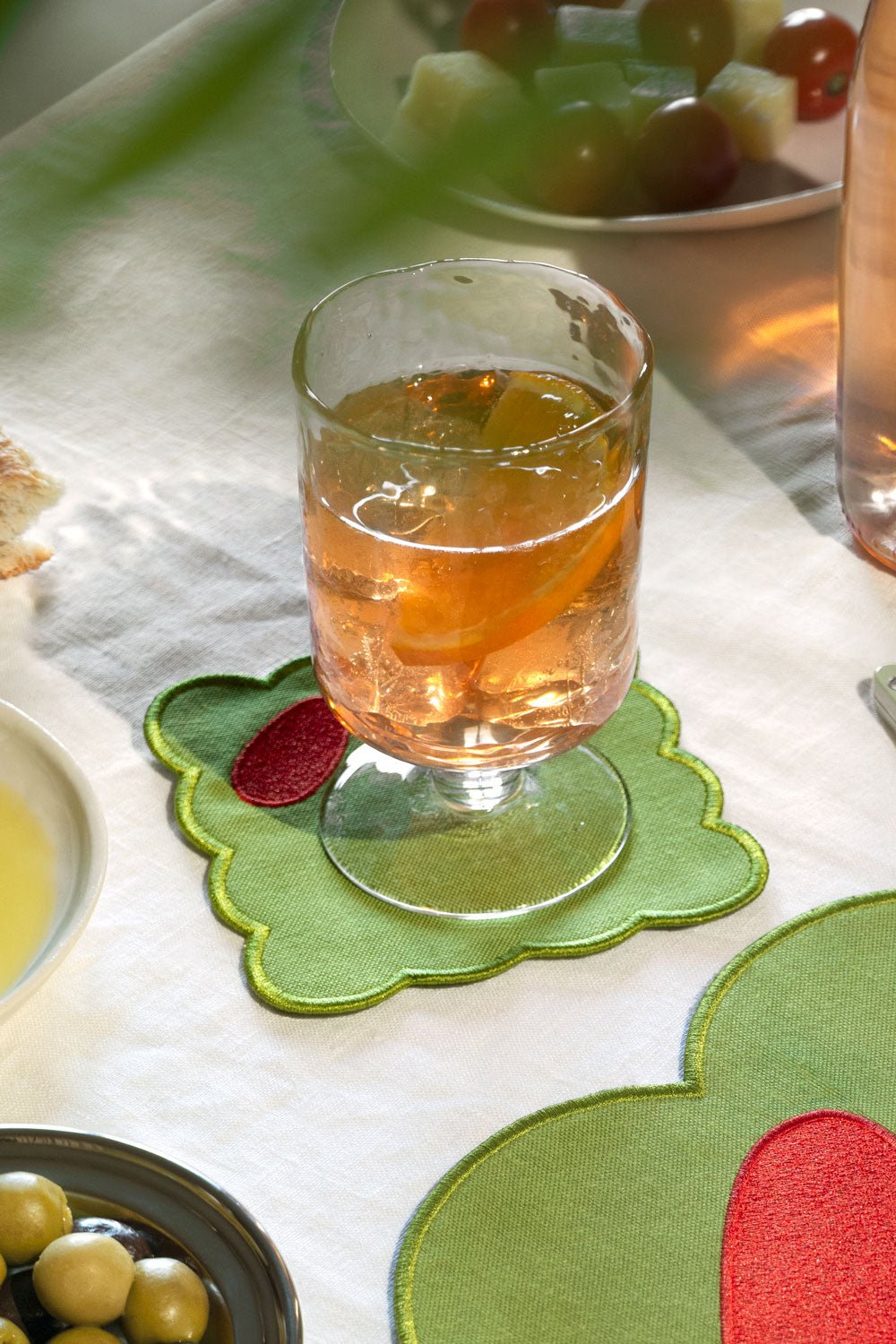 A glass filled with an drink sits on a green olive fabric coaster, placed on a white tablecloth in a dining setting.
