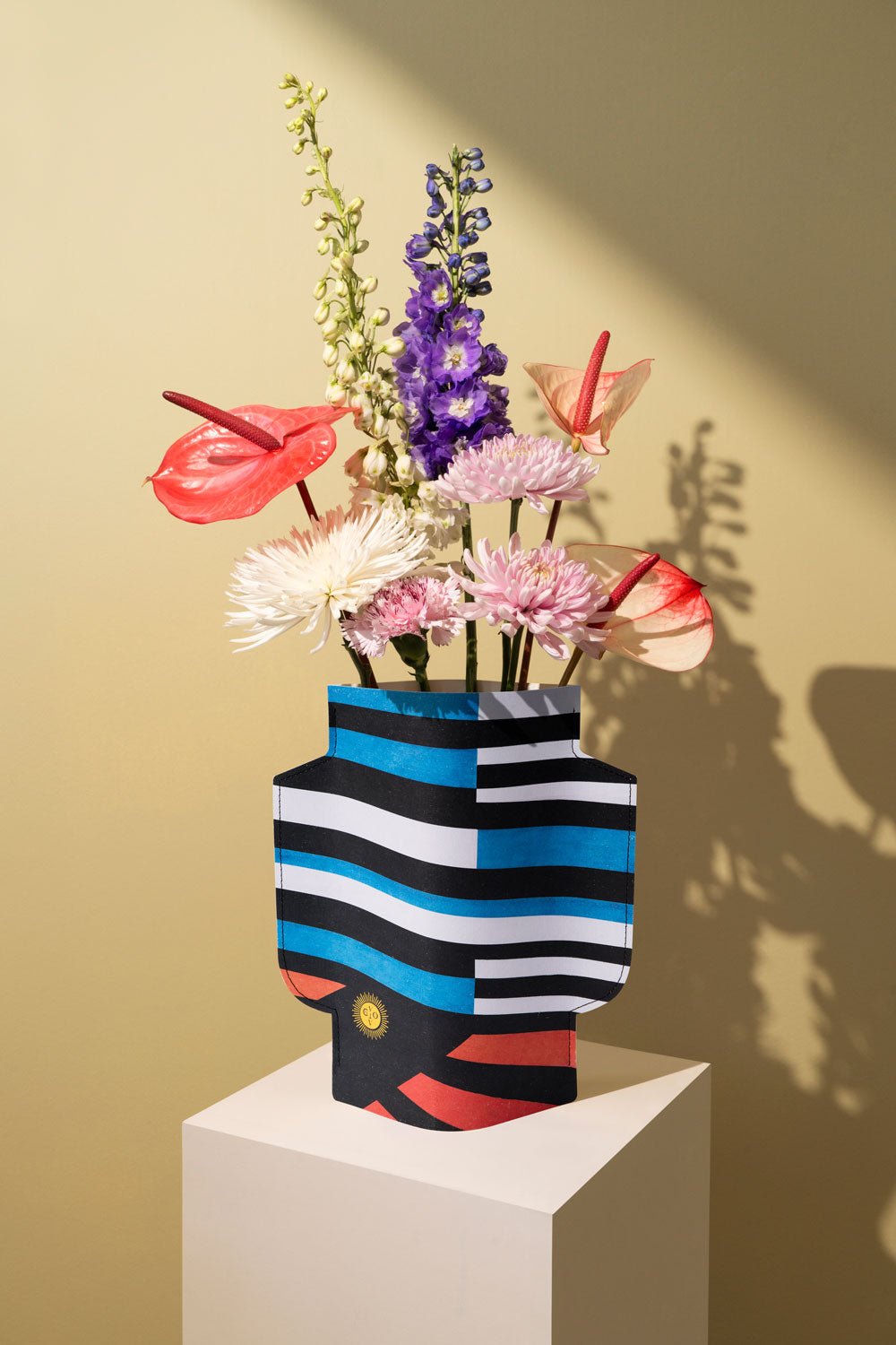 A vibrant bouquet fills the OCTAEVO Palma Paper Vase, displayed on a pedestal with striking lighting and a beige backdrop.