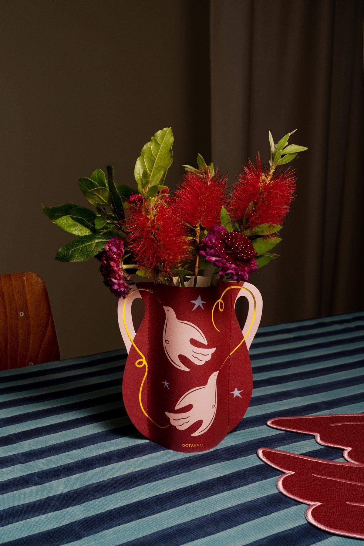 Le mini vase en papier Paloma OCTAEVO, orné de motifs de colombes, est posé sur une nappe à rayures bleues et accueille des fleurs.