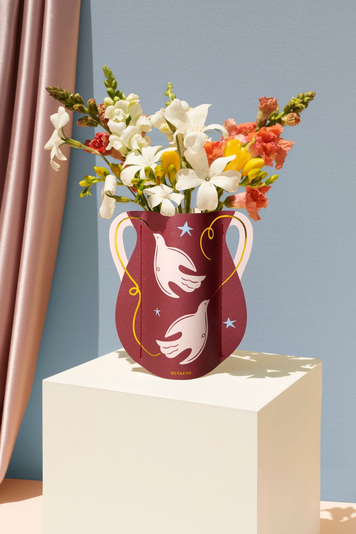The Paloma Mini Paper Vase by OCTAEVO with white bird motifs displays blooms on a pedestal and pastel backdrop.