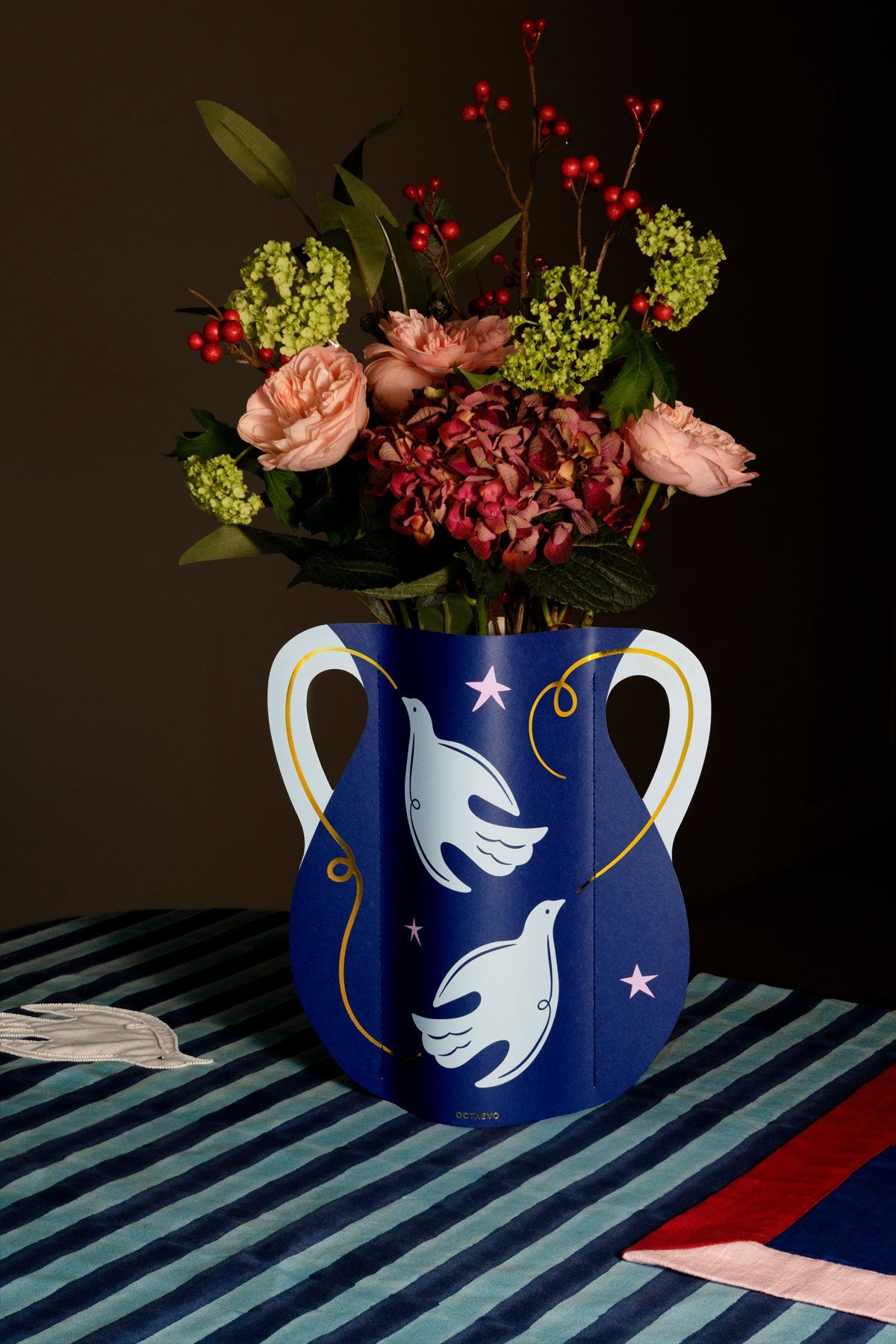 Le vase en papier Paloma OCTAEVO, garni de fleurs roses, est posé sur des rayures, à côté d'un masque et d'une serviette pliée.
