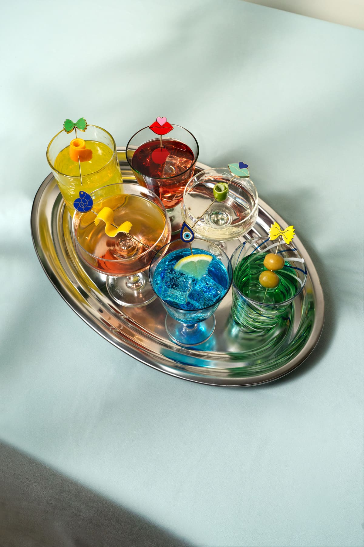Six cocktails on a silver tray, each garnished with a different OCTAEVO gold-plated cocktail pick — farfalle, lips, evil eye and more.