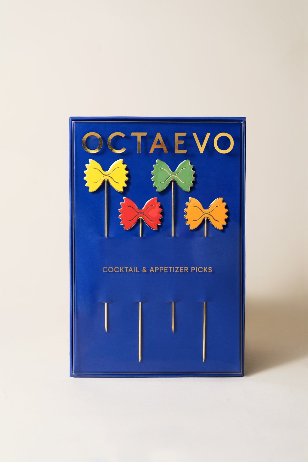 A blue OCTAEVO box holds four gold-plated cocktail picks shaped like bow-tie pasta—Pasta Party set.
