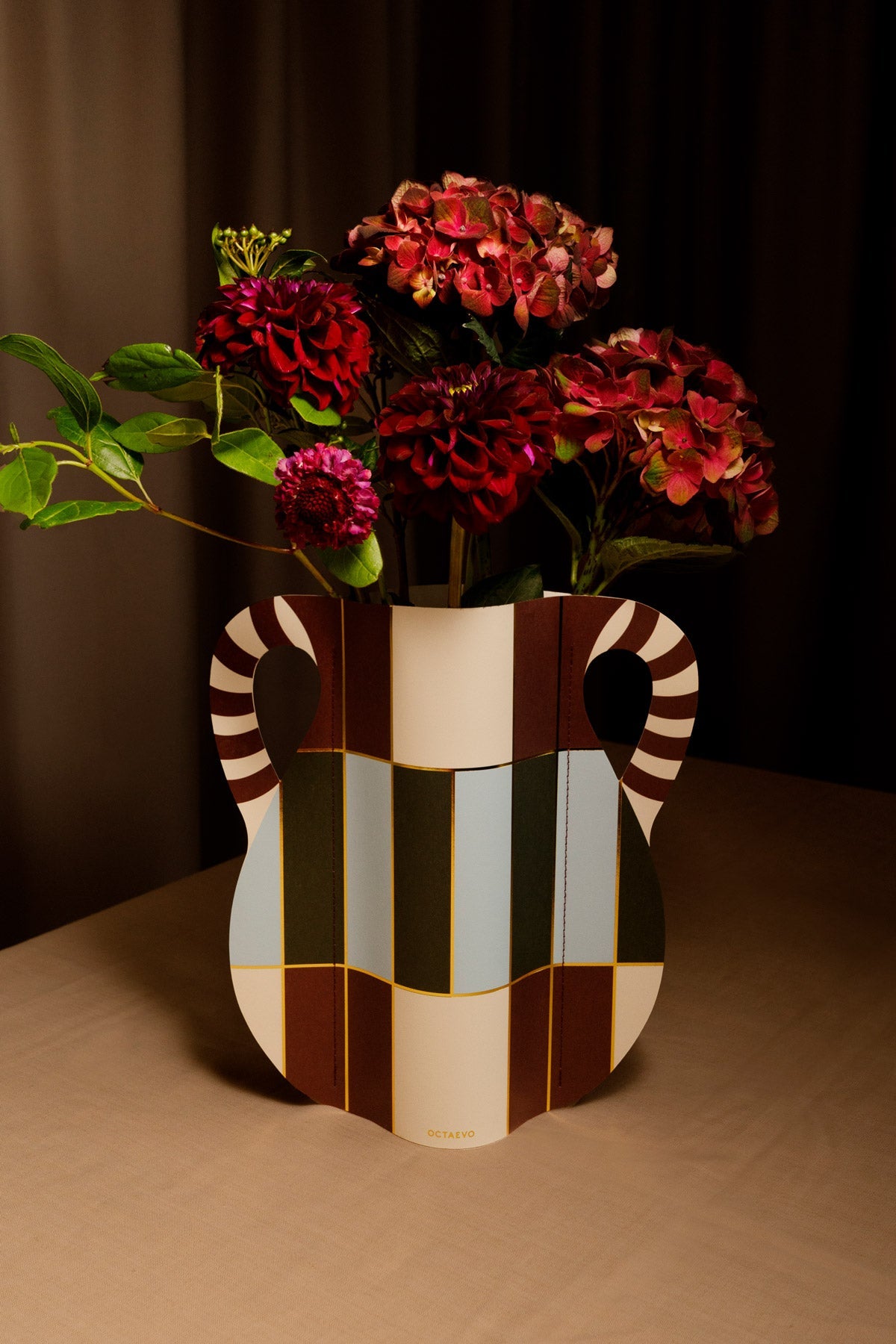 Rote Blumen in OCTAEVOs bunter Patio Papiervase mit gestreiften Henkeln auf einem beigen Tisch neben einem dunklen Vorhang.
