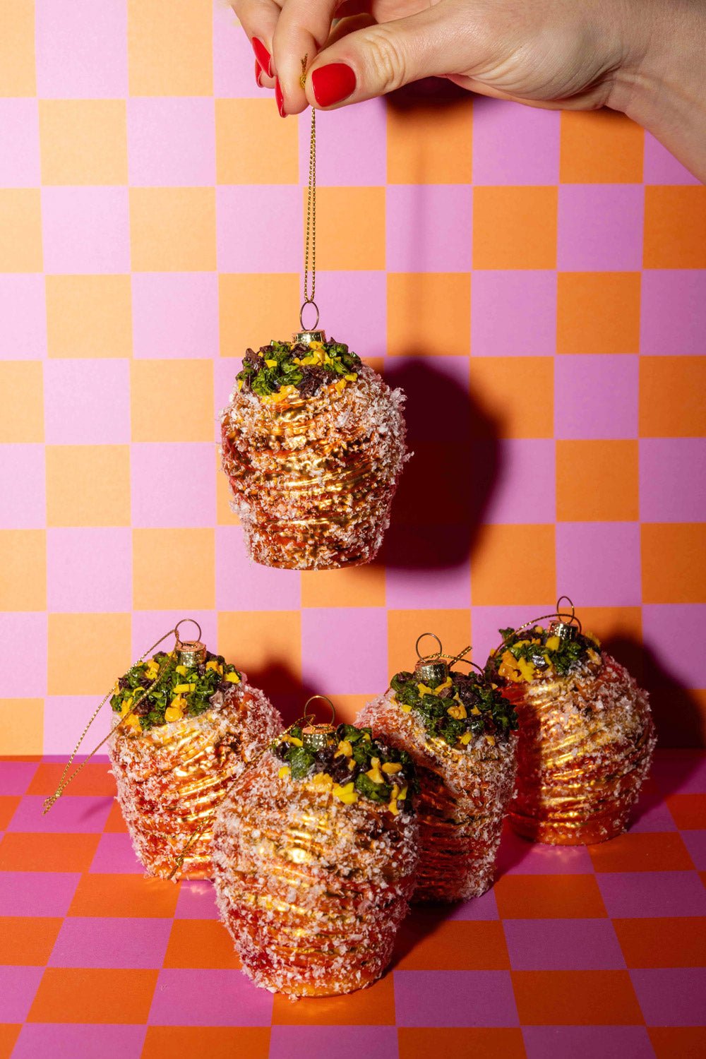 A hand holds the OCTAEVO Pistachio Cruffin Ornament above four others on a pink and orange checkered surface.