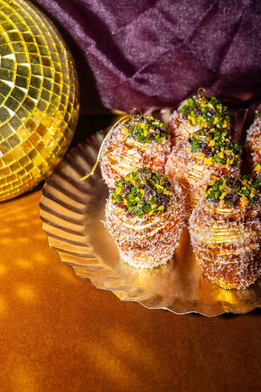 Seis Adornos Cruffin de Pistacho de OCTAEVO añaden encanto festivo a un plato dorado, junto a una bola de discoteca amarilla y una tela morada.
