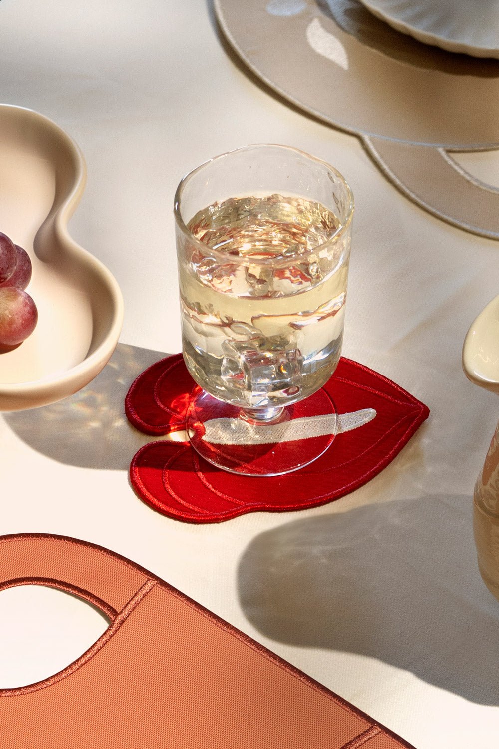 Un verre de vin blanc sur les dessous de verre Anthurium rouges d'OCTAEVO (lot de 4), à côté de raisins et d'arts de la table élégants.