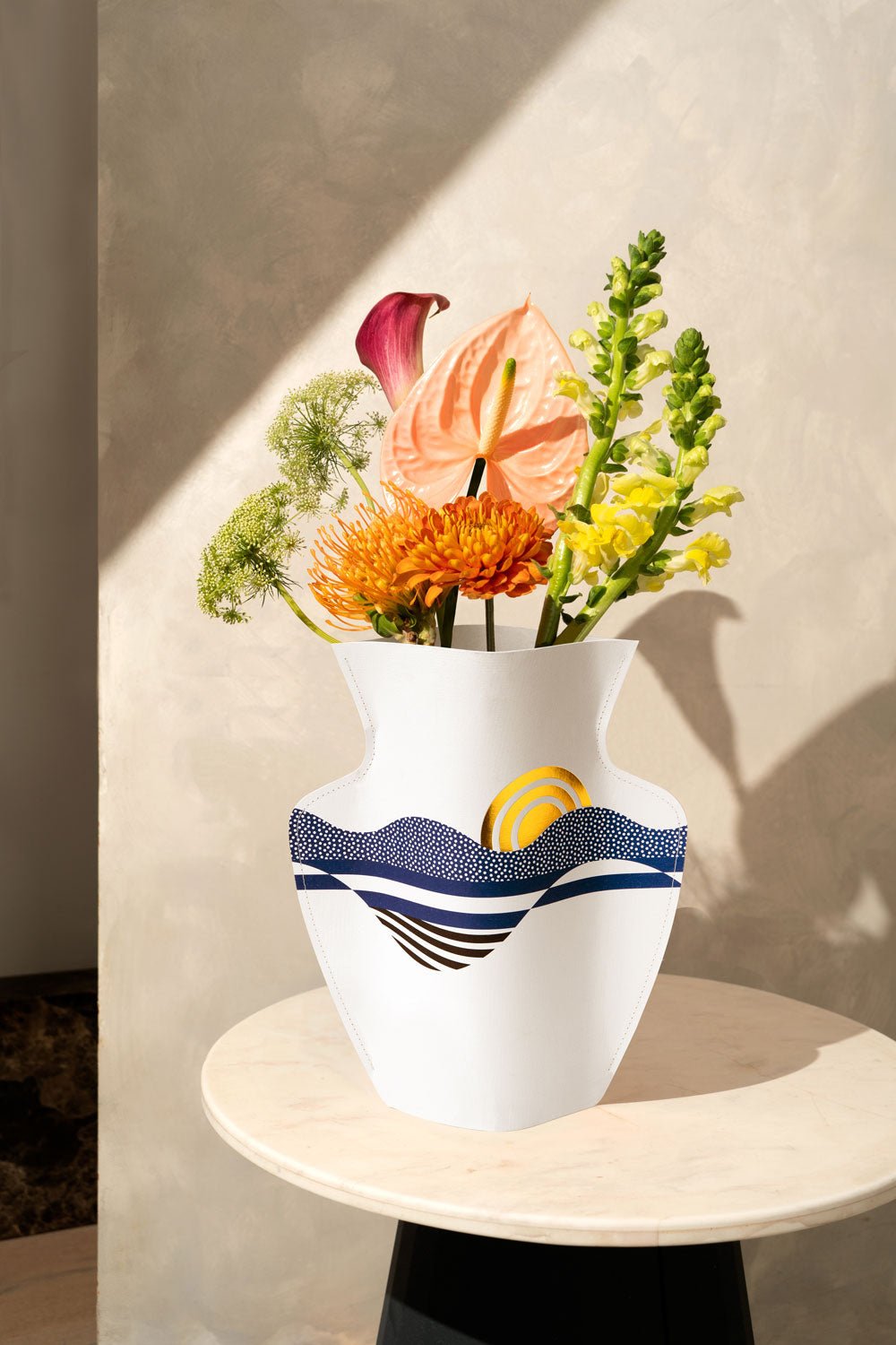The Salina Paper Vase by OCTAEVO with blue and yellow patterns holds blooms on a sunlit round table.