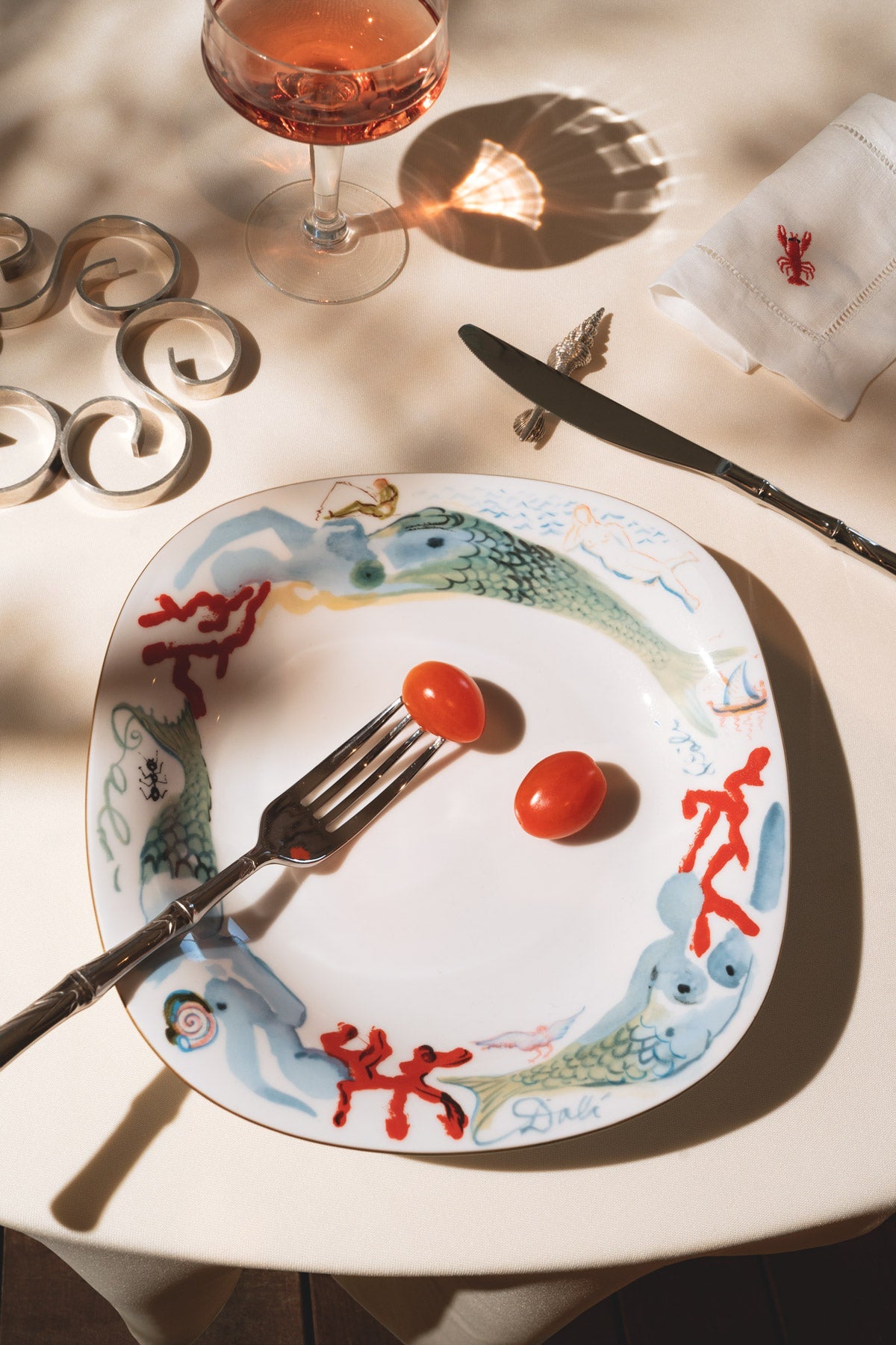 Une fourchette, des tomates cerises sur une assiette Sirène Salvador Dalí OCTAEVO, un couteau, une serviette et un rosé, sur une table ensoleillée.