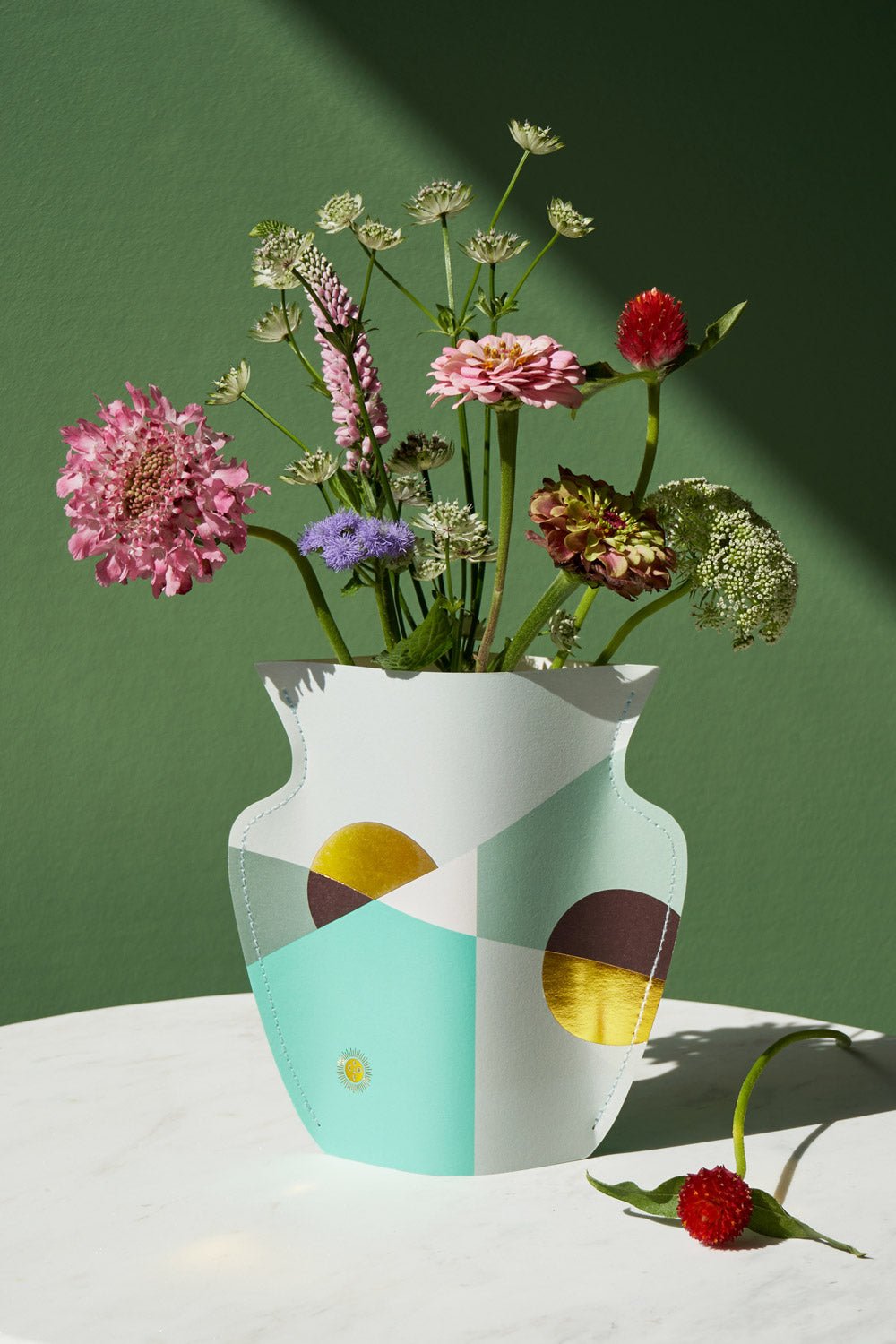 Colorful wildflowers in the OCTAEVO Siena Mini Paper Vase on white; a red bloom lies nearby against a green background.