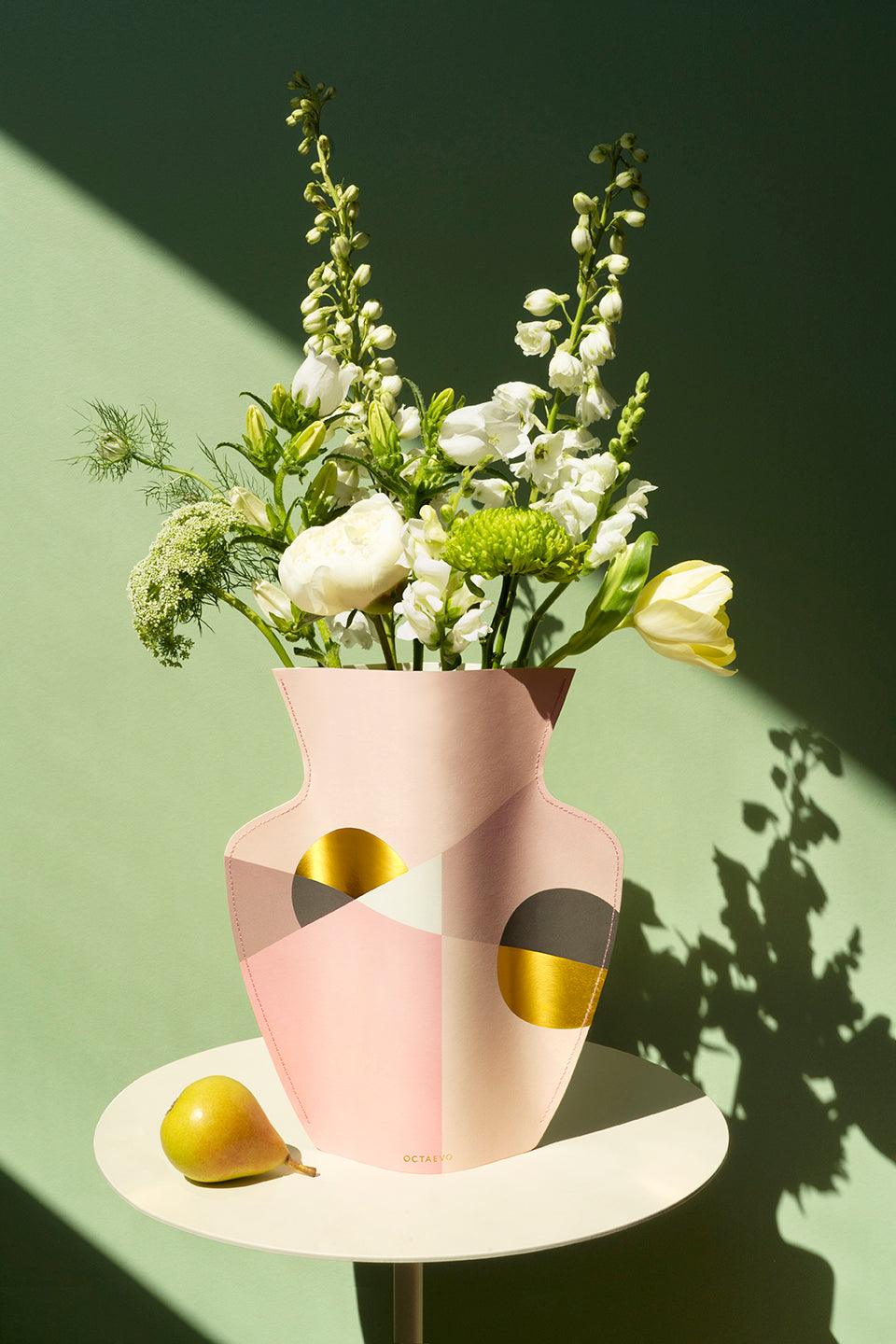 The pink Siena Paper Vase by OCTAEVO with gold accents holds blooms on a round table beside a pear.