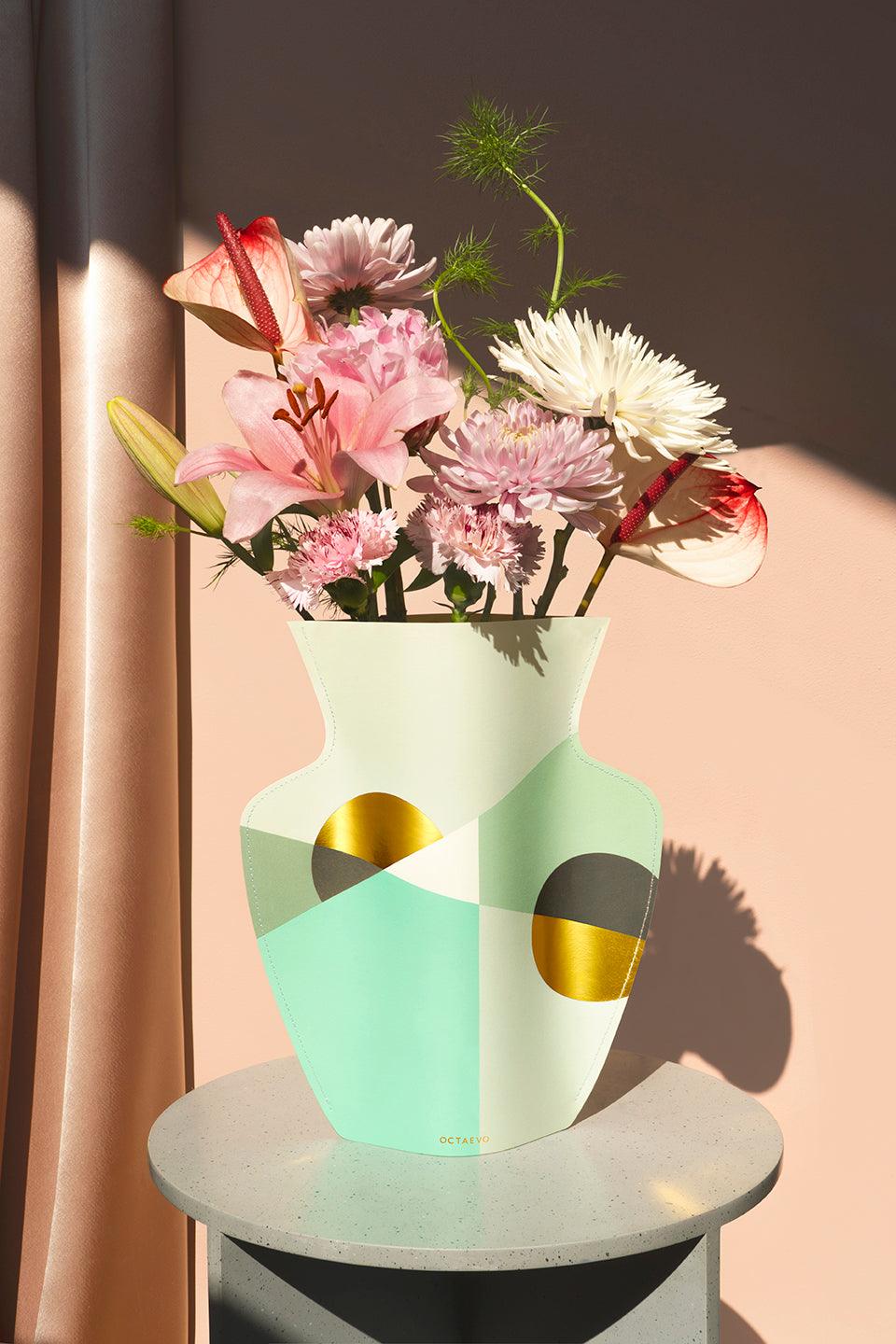 The mint OCTAEVO Siena Paper Vase with pastel blooms on a round gray table against a peach background.