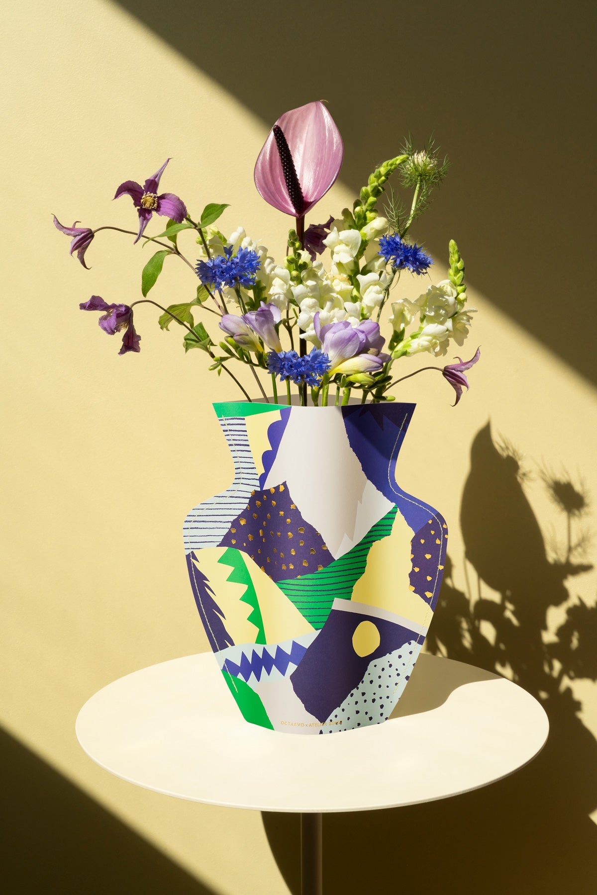 The OCTAEVO Stromboli Paper Vase holds assorted flowers on a round white table against a yellow wall.