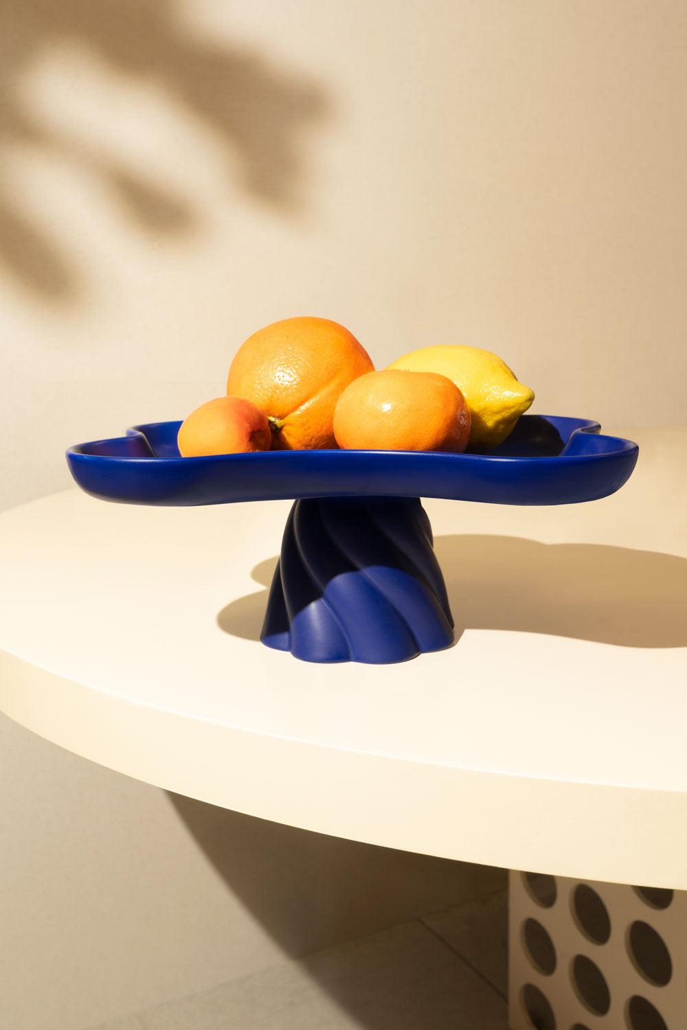 A blue Octaevo Templo High Tray displays oranges, a lemon, and an apricot on a white table with a patterned base.