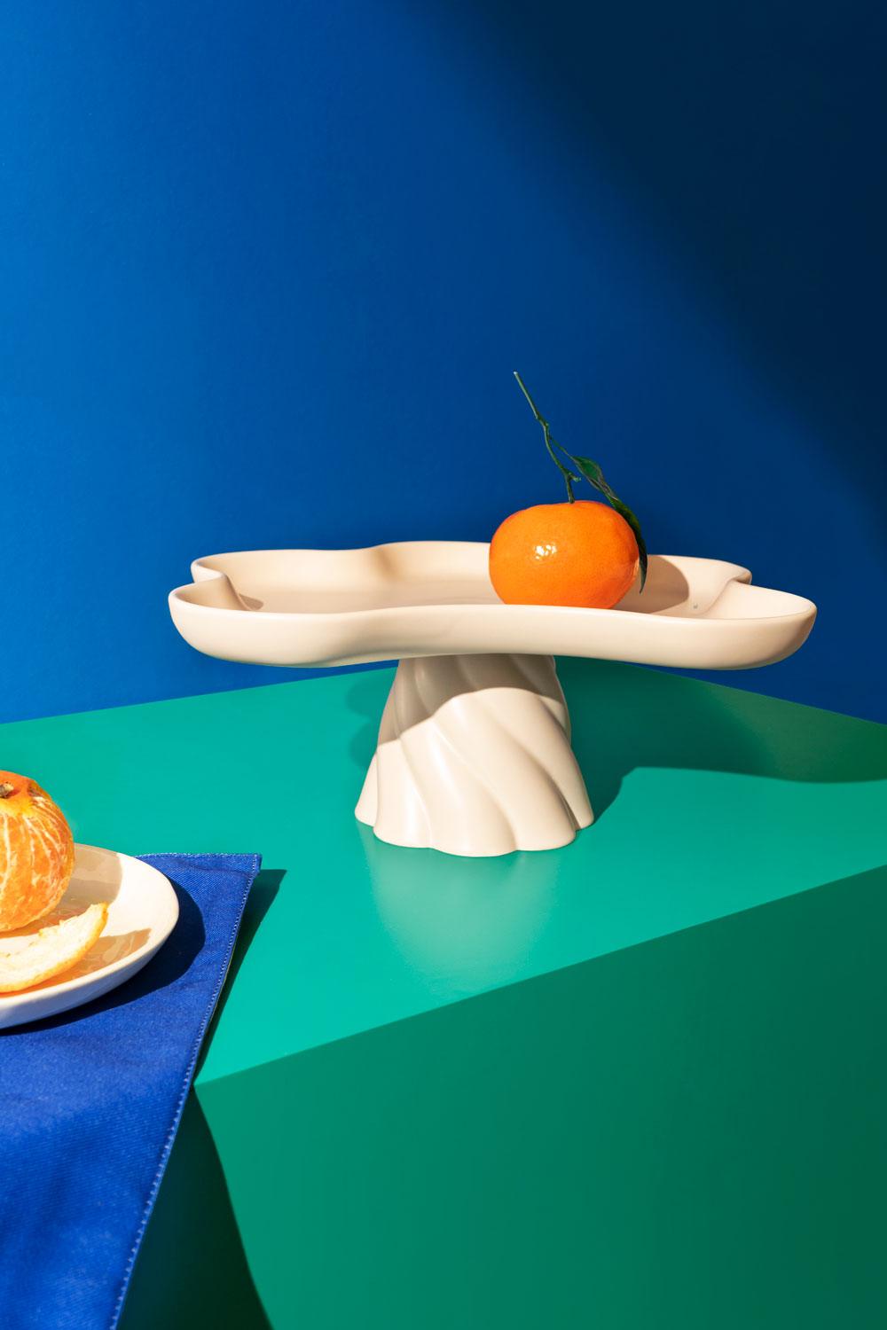 An orange with a leaf sits on an Octaevo Templo High Tray in ivory beside sliced oranges on a blue-green surface.