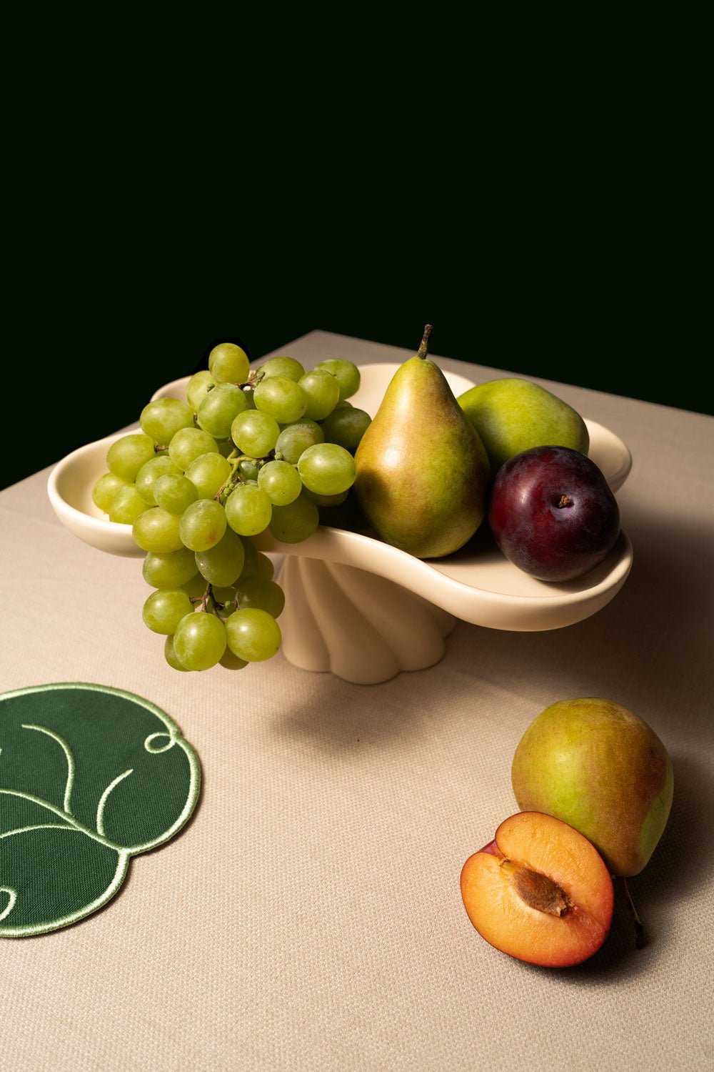 An ivory OCTAEVO Templo High Tray filled with green grapes, a pear, and a plum, on a beige tablecloth with a coaster nearby