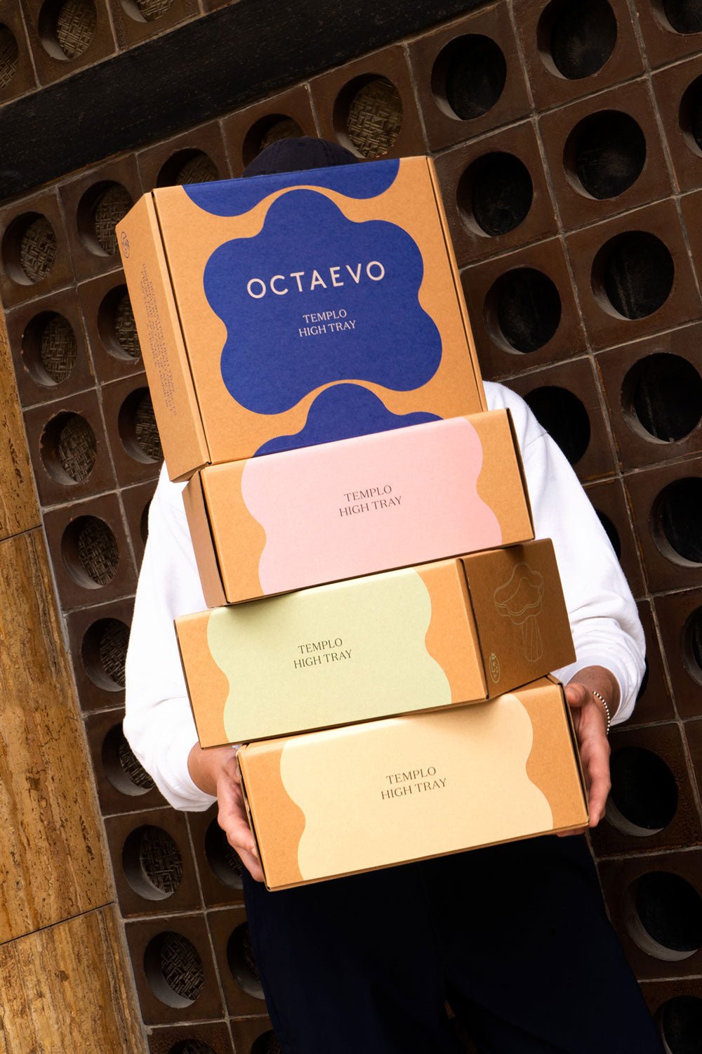 Person holds four stacked OCTAEVO Templo High Tray boxes in front of a brown patterned wall.