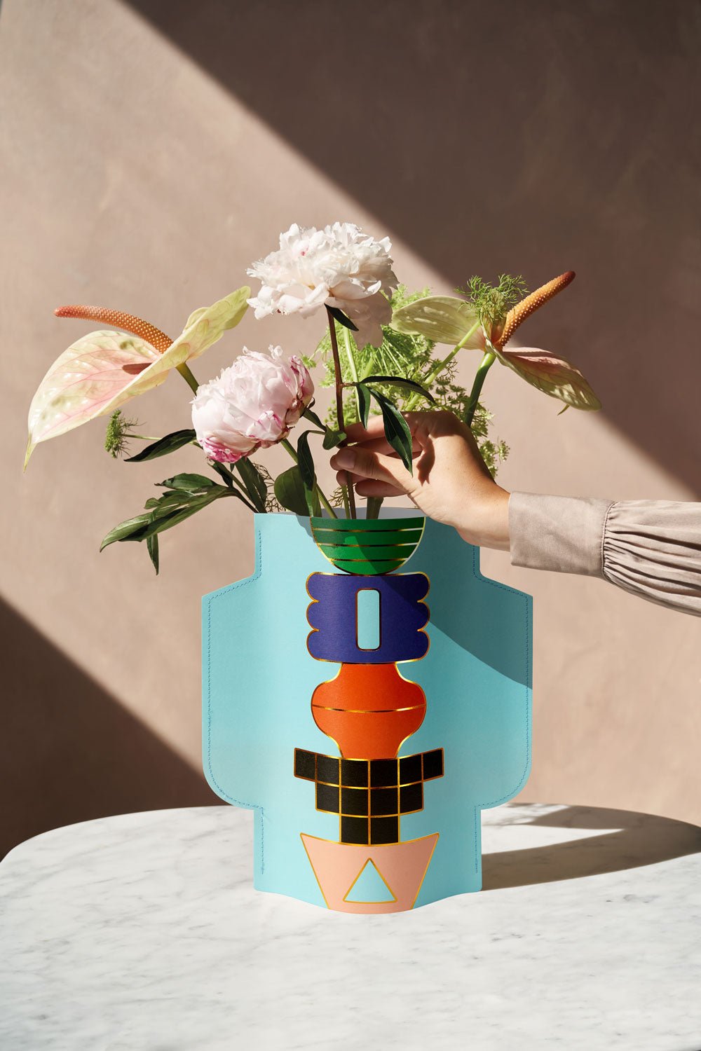 A light blue OCTAEVO Paper Vase with a geometric design, filled with white and pink flowers. A hand is adjusting the flowers.