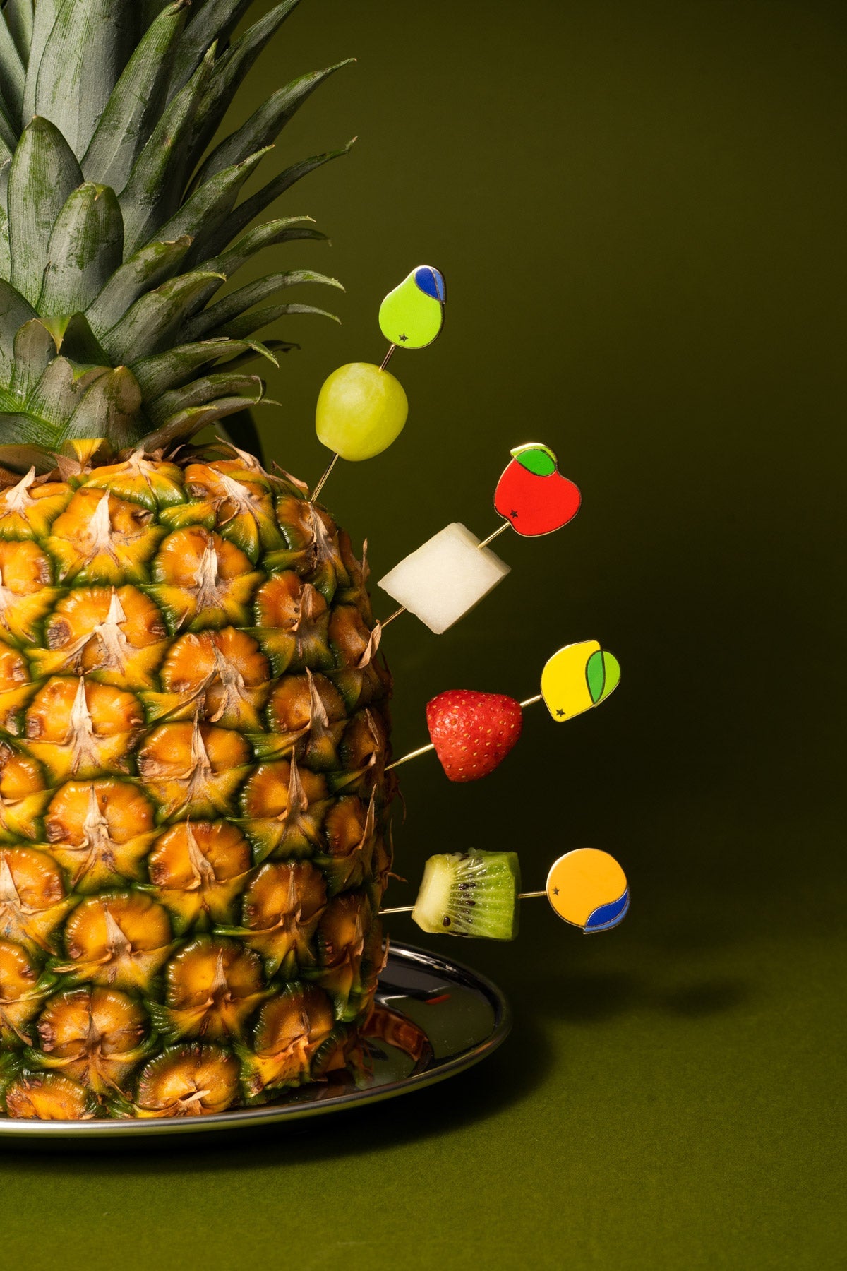Octaevo’s Tutti Frutti Cocktail Picks (Set of 4) hold vibrant fruits, styled against a bold green background.