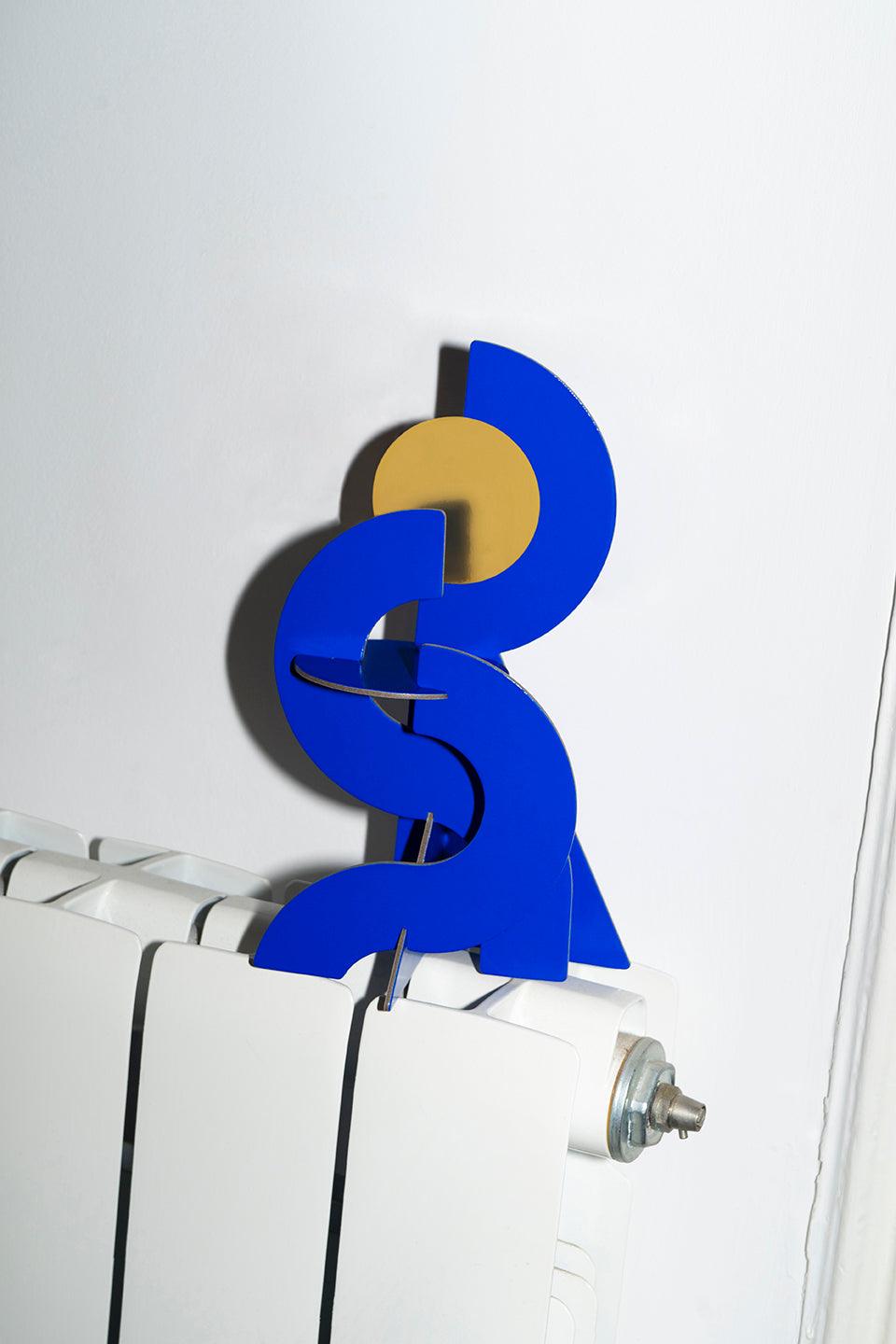 Octaevos Water Paper Sculpture, in abstract blue and gold, sits on a white radiator against a white wall.