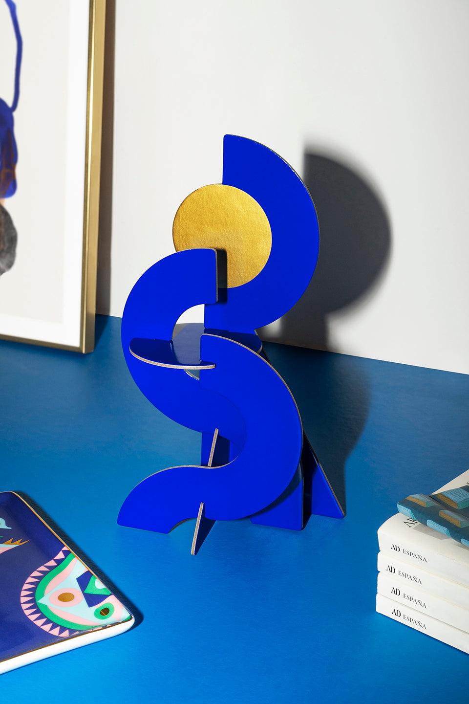 The Water Paper Sculpture by Octaevo, in blue and gold, adds modern art style to your home decor on a blue desk.
