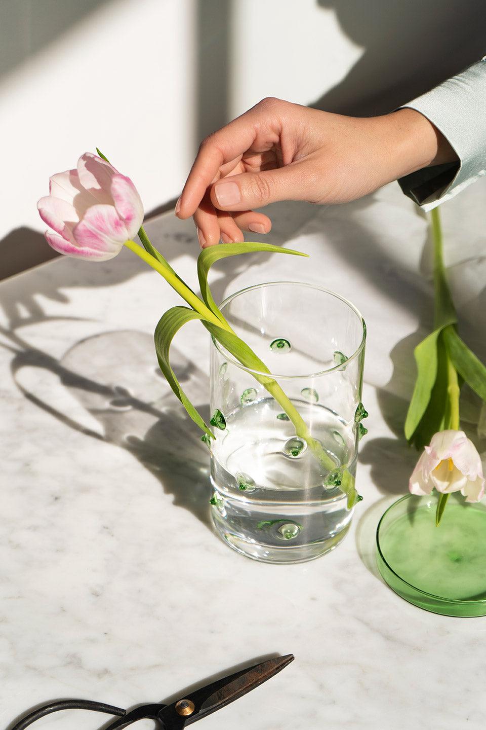 Eine Hand stellt eine rosa Tulpe in den Agave Glasbehälter Groß von OCTAEVO auf einem Marmortisch, daneben eine Schere.