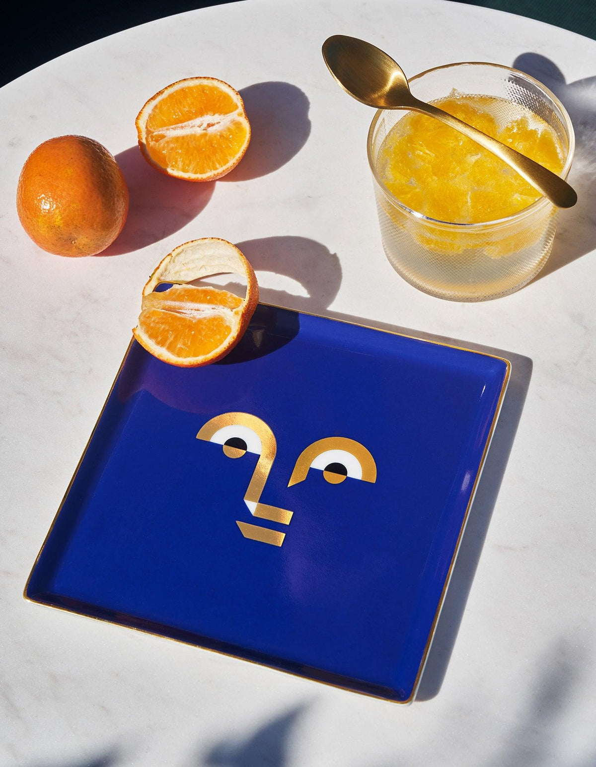 The OCTAEVO Apollo Ceramic Tray in blue with a gold face design, shown beside oranges and glass.
