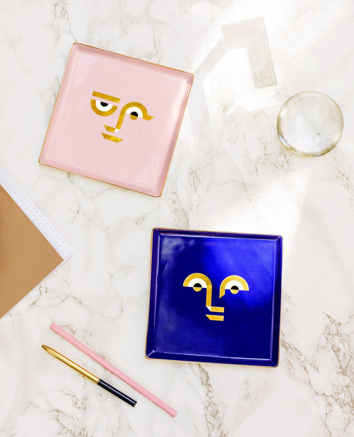 Two OCTAEVO Apollo Ceramic Trays, pink and blue, with abstract gold faces, shown on marble with pens and a notebook.