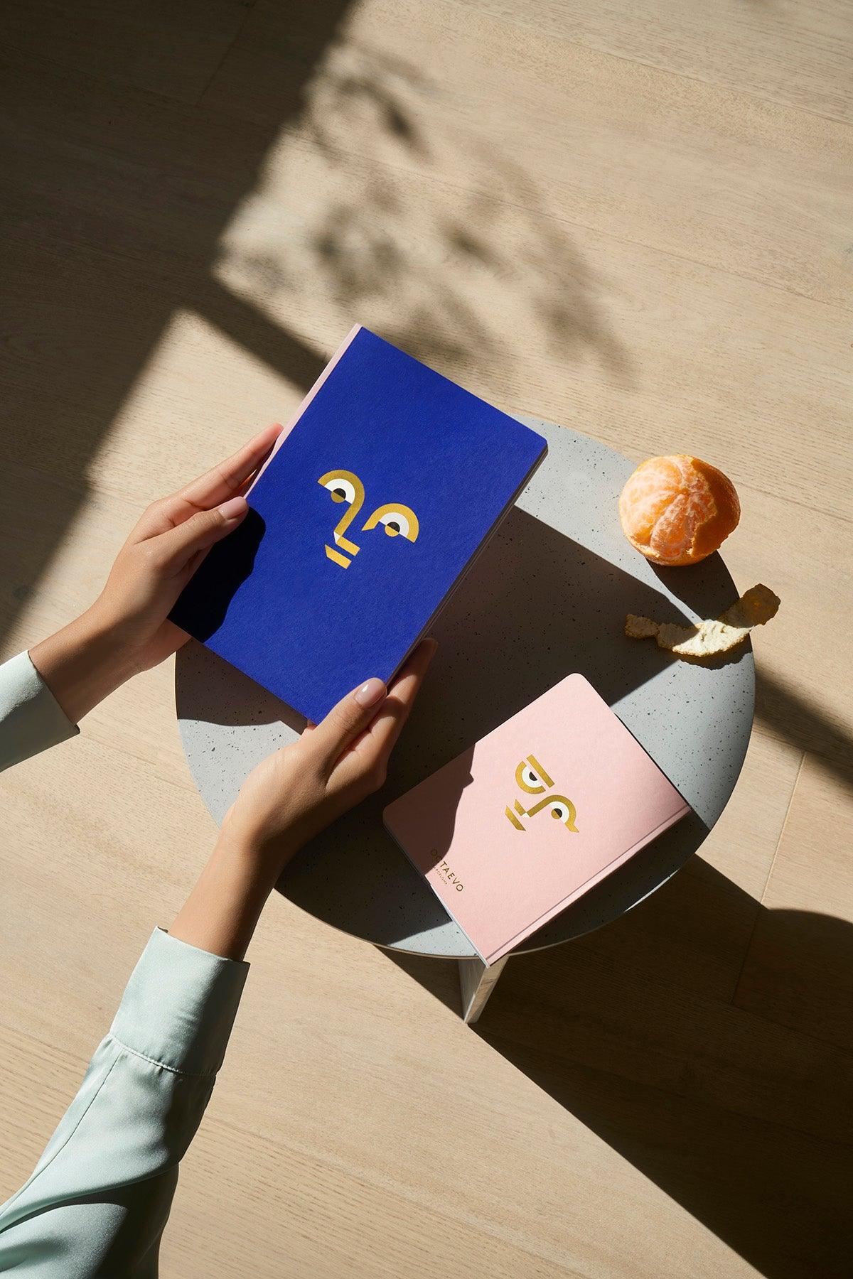A person holds an Octaevo Apollo Notes A5 notebook beside a pink notebook and a peeled orange on a table.