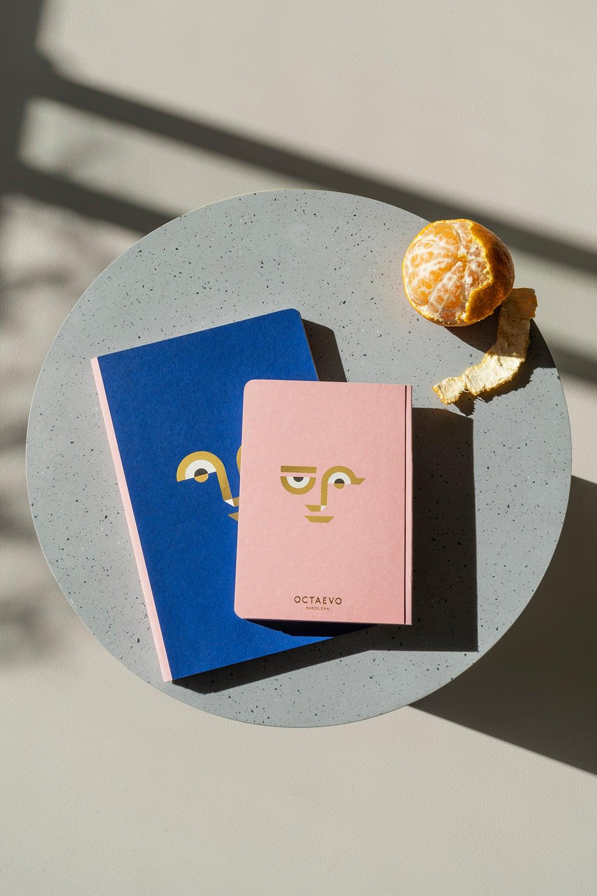 Two Octaevo Apollo Notes A6 notebooks with lay flat binding and a peeled orange on a sunlit round gray table.
