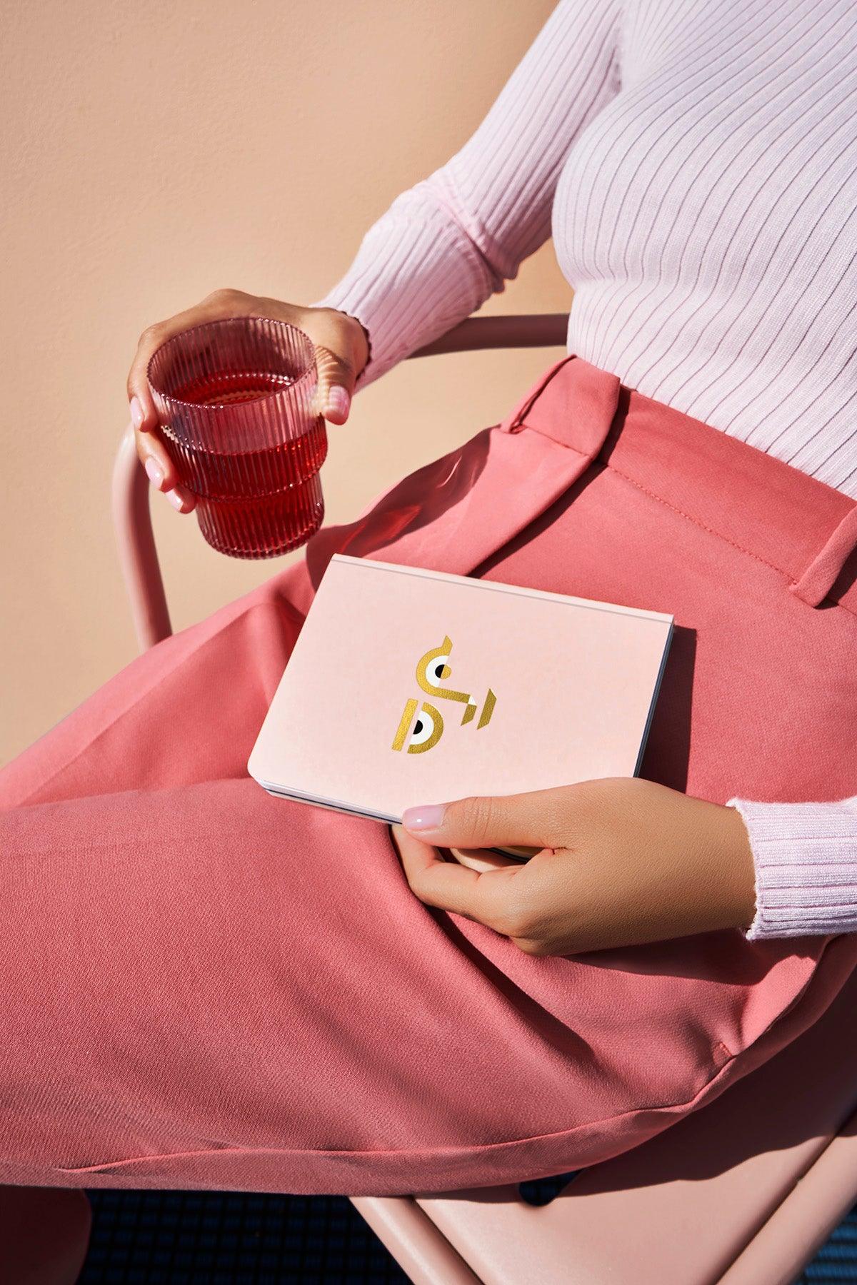Woman in pink skirt holds a red drink and an OCTAEVO Apollo Notebook A6, seated on a pink chair.
