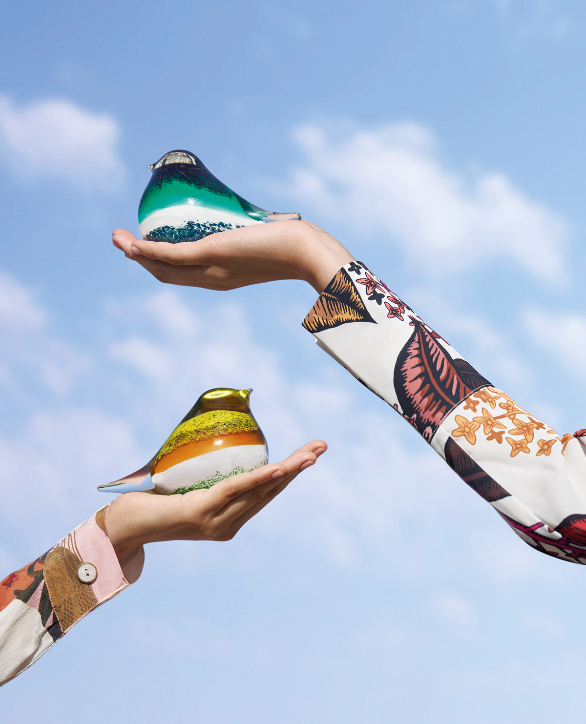 Two hands holding colorful glass bird figurines against a blue sky, wearing patterned long sleeve shirts.