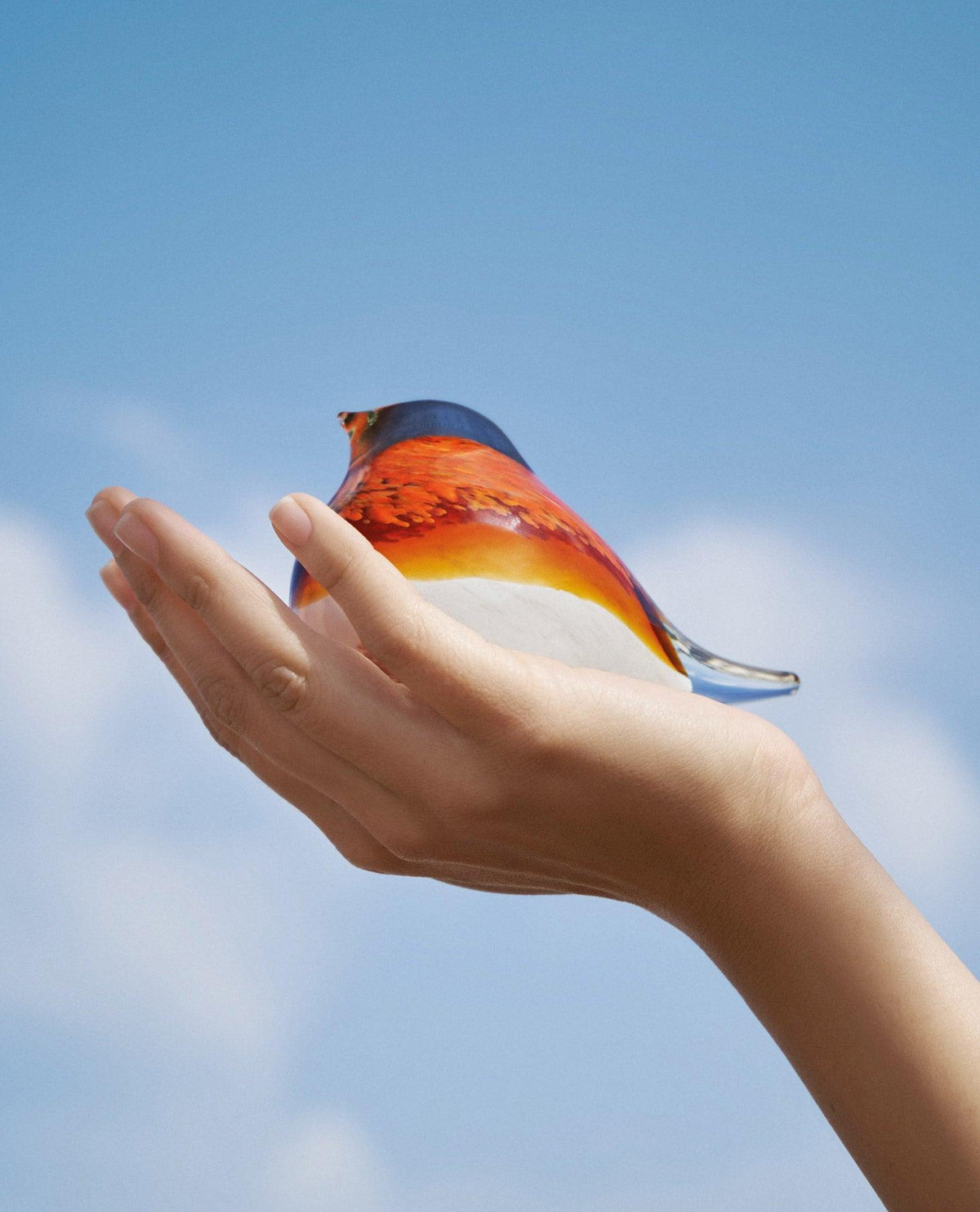 A hand gently holds an orange OCTAEVO Atlas Glass Bird, a vibrant statement piece, against a blue sky.