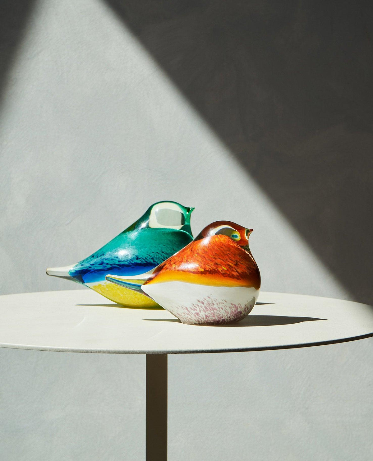Colorful OCTAEVO Atlas Glass Bird sculptures sit on a round table, casting shadows under dramatic lighting.