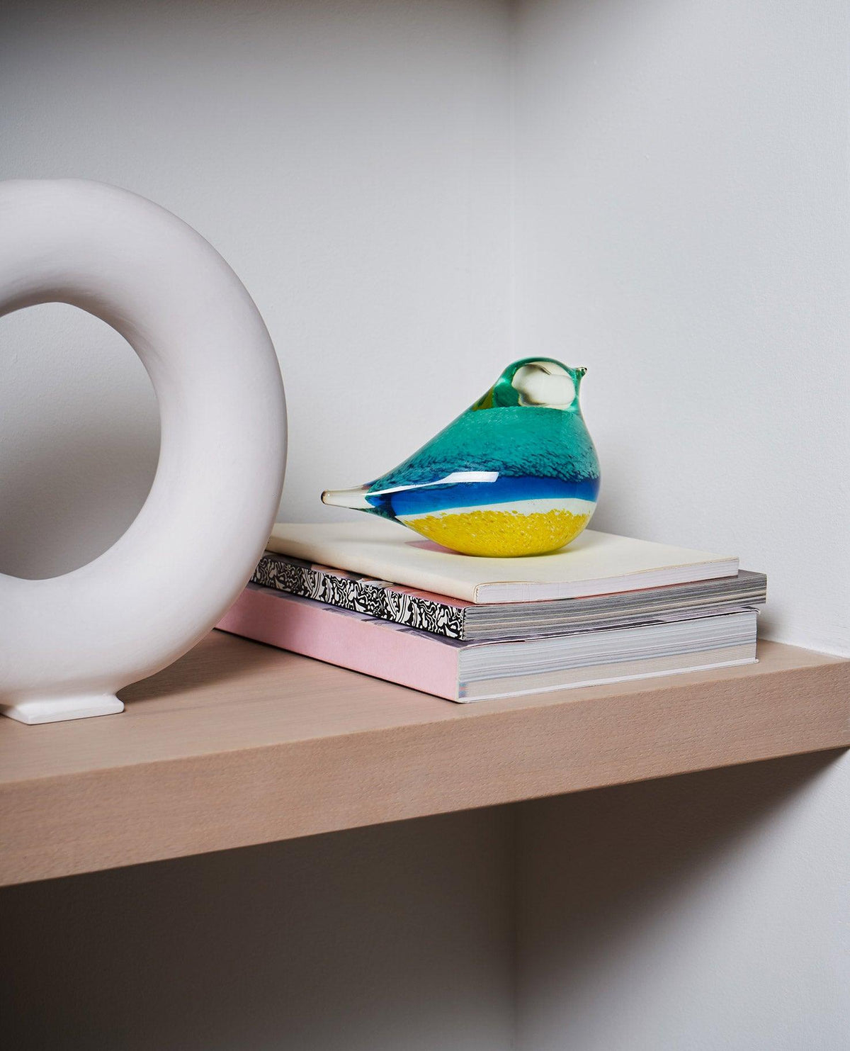 The blue and yellow OCTAEVO Atlas Glass Bird sits on stacked books beside a white circular sculpture on a wooden shelf.