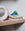 The blue and yellow OCTAEVO Atlas Glass Bird sits on stacked books beside a white circular sculpture on a wooden shelf.