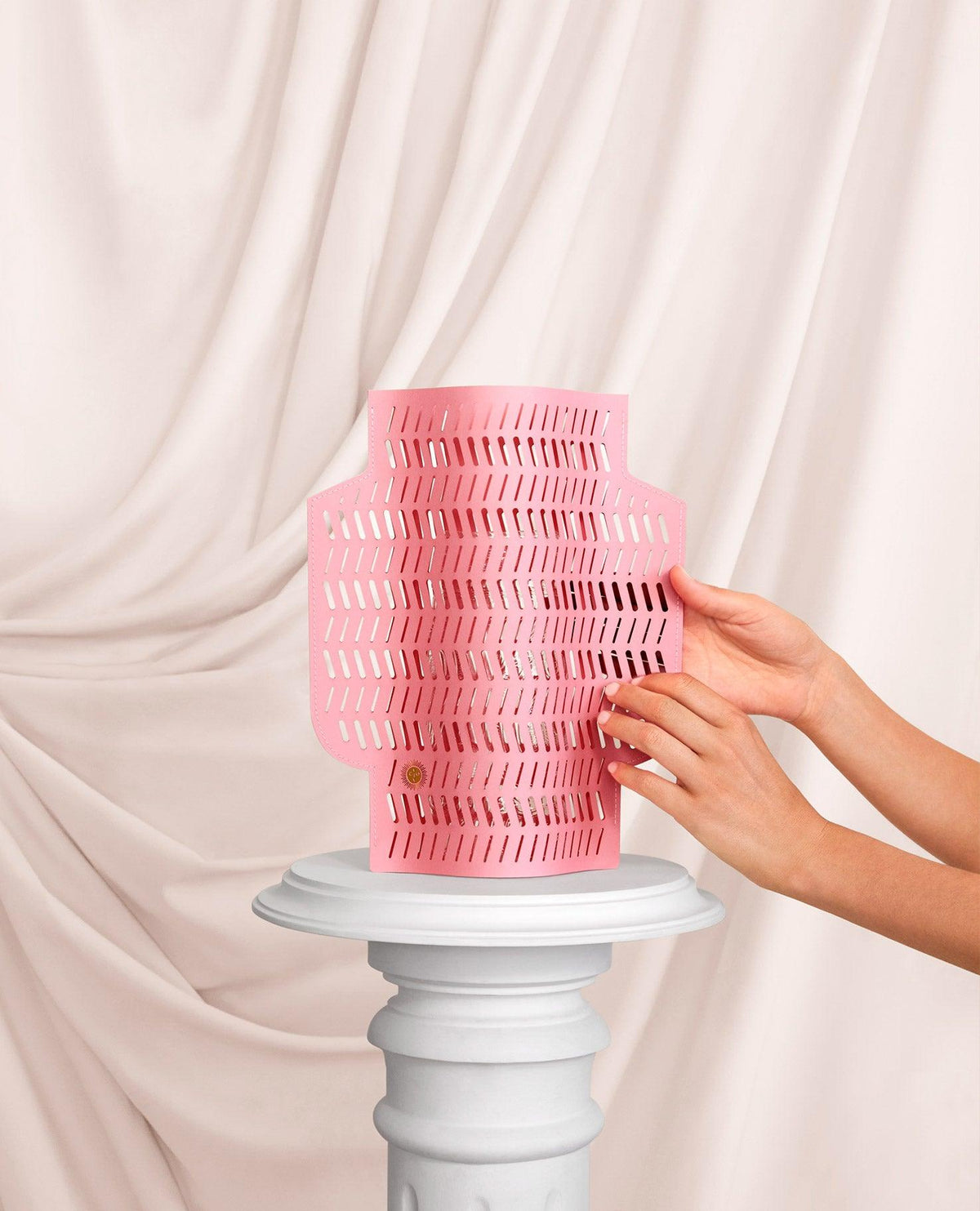 Deux mains tiennent le vase en papier Aurea par OCTAEVO au-dessus d'un piédestal blanc, avec un tissu crème drapé en arrière-plan.