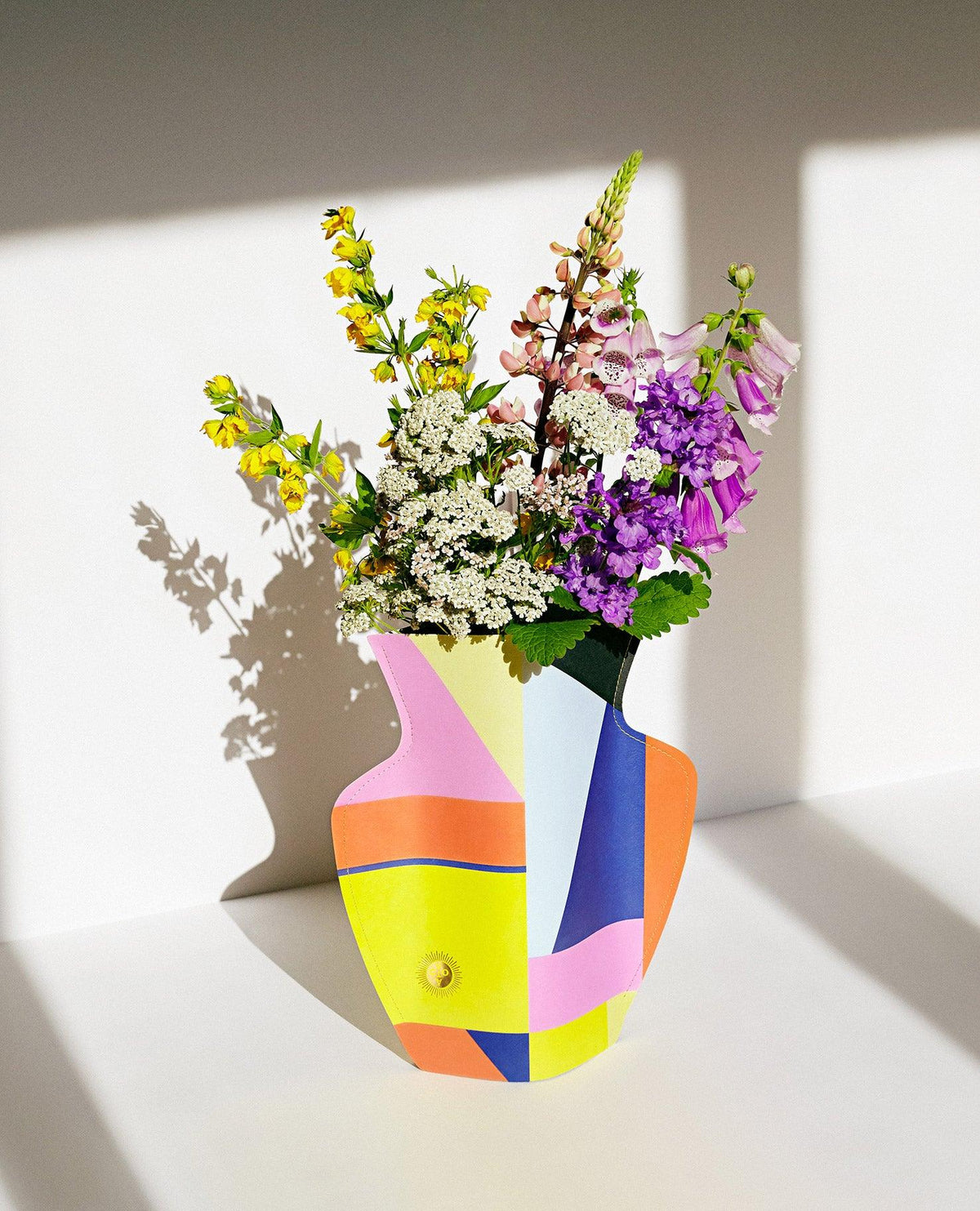 The OCTAEVO Bazaar Paper Vase displays vivid wildflowers, casting a shadow on a white surface in bright sunlight.