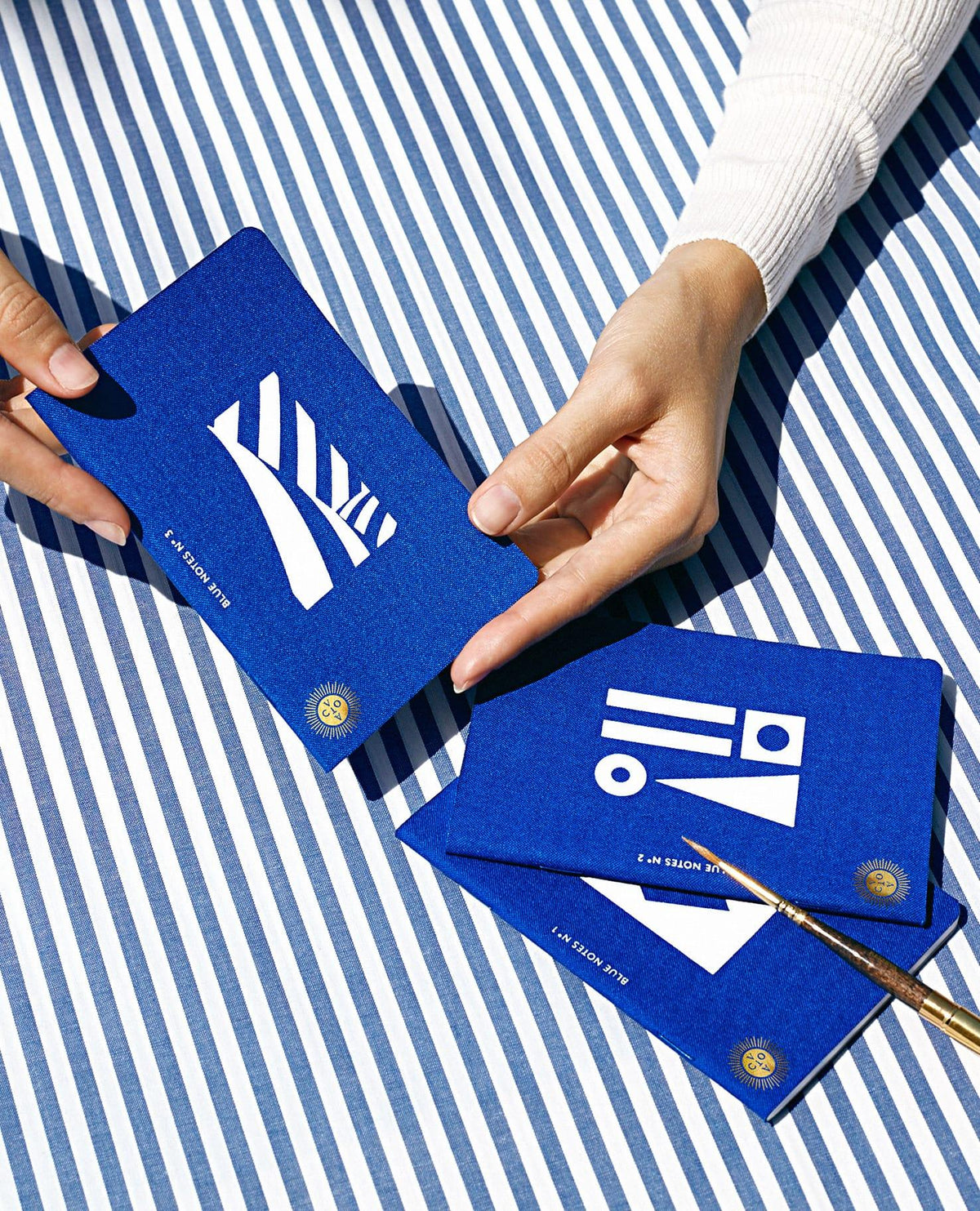 Des mains tiennent les carnets Passport bleus d'OCTAEVO au-dessus de rayures, avec des formes blanches sur une couverture bleue éclatante.