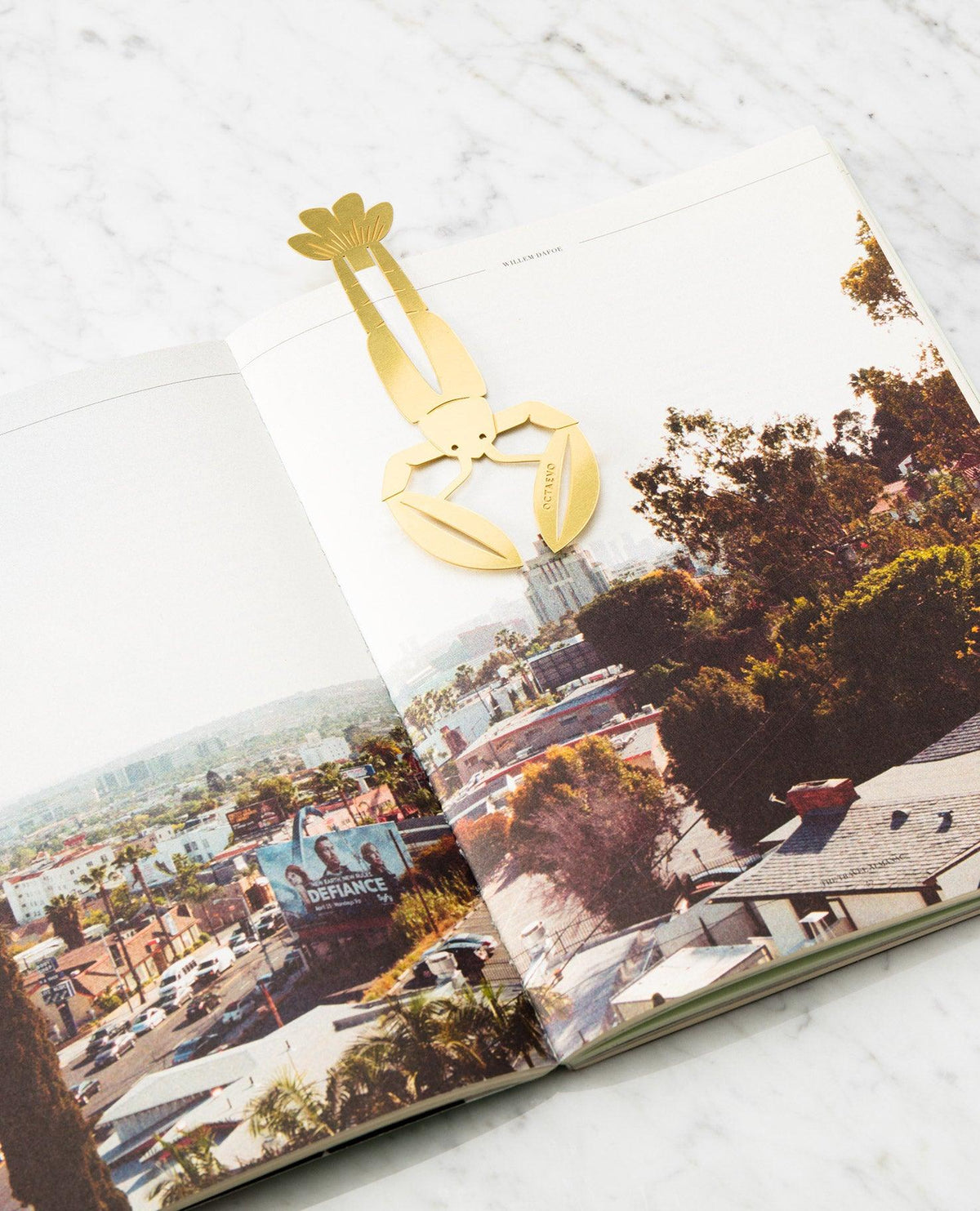 The OCTAEVO Bookmark Bon Vivant, a gold brass lobster bookmark, rests on an open book—a unique gift idea.