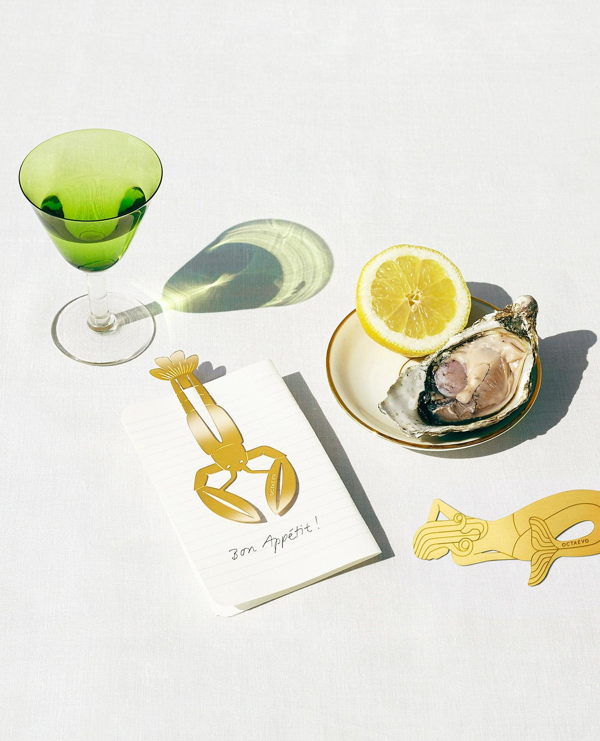 A gold bon Vivant lobster and mermaid bookmark by OCTAEVO, green glass, oyster with lemon, on a white tablecloth.