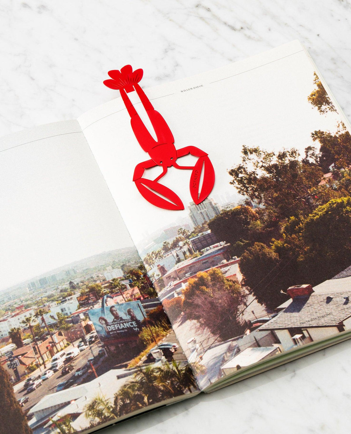 The OCTAEVO Bookmark Bon Vivant in red brass rests on an open book with a striking cityscape photo.
