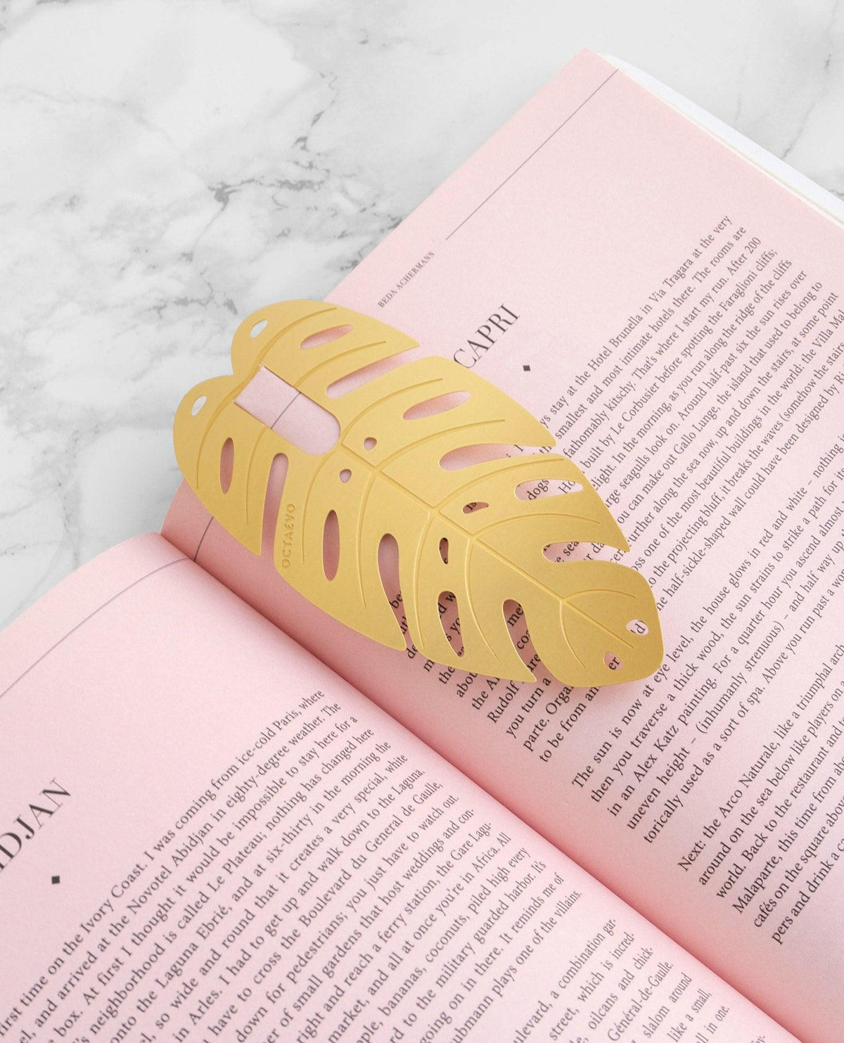 The Bookmark Capri by OCTAEVO, a brass monstera leaf, rests on an open pink book atop a marble surface.