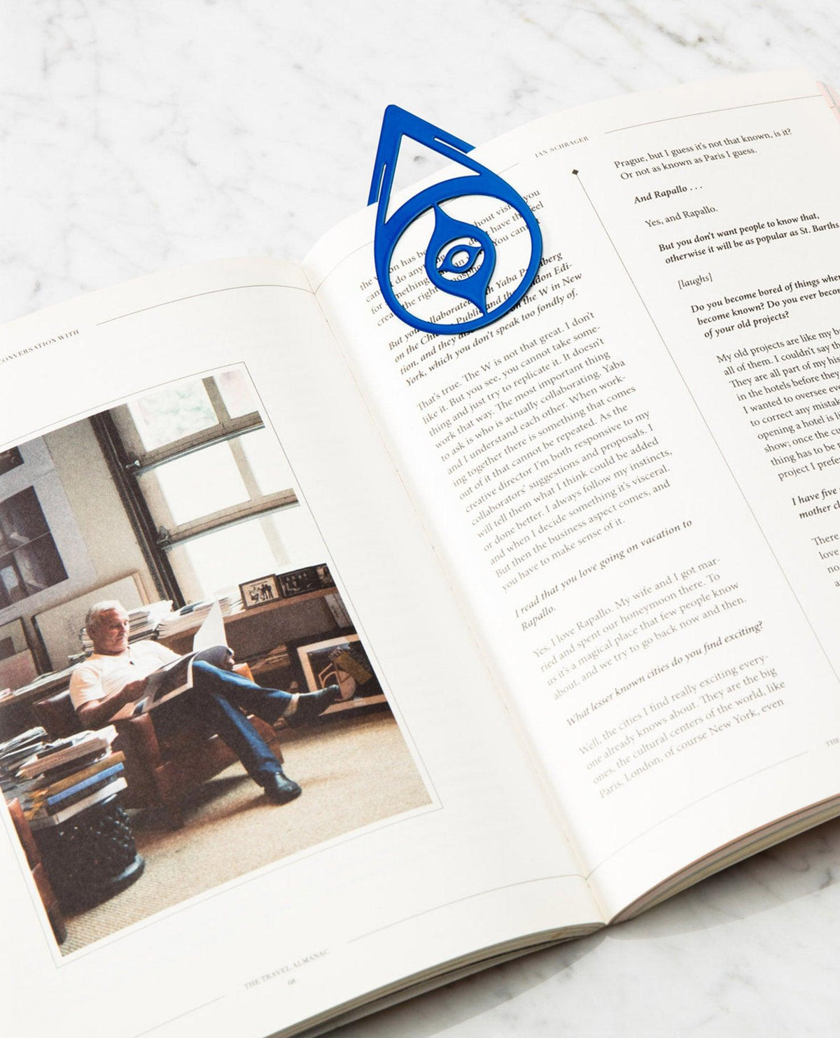 Bookmark Nazar by OCTAEVO: open book on marble, brass blue bookmark clipped on page. 