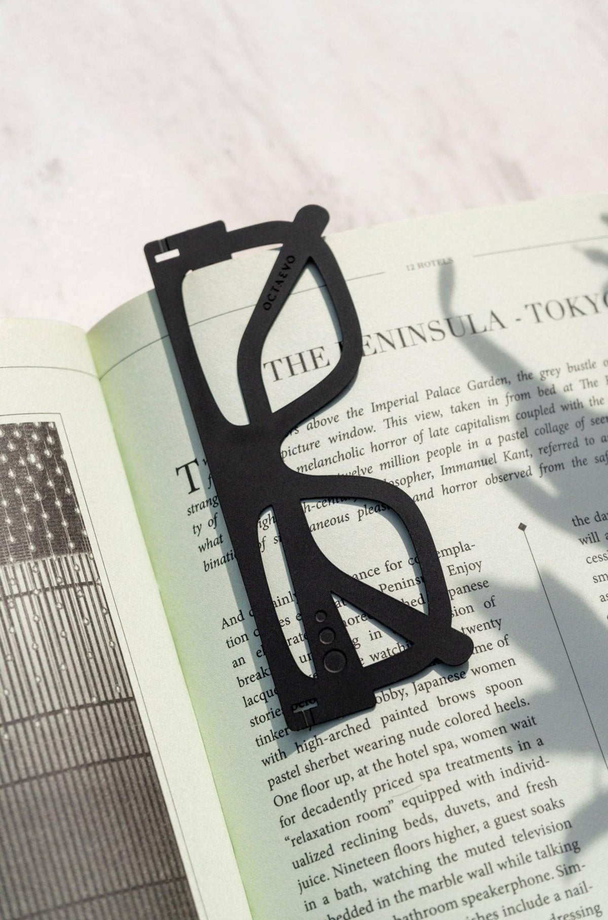 The black OCTAEVO Bookmark Plein Soleil, shaped like sunglasses, sits on an open book with text and an illustration.