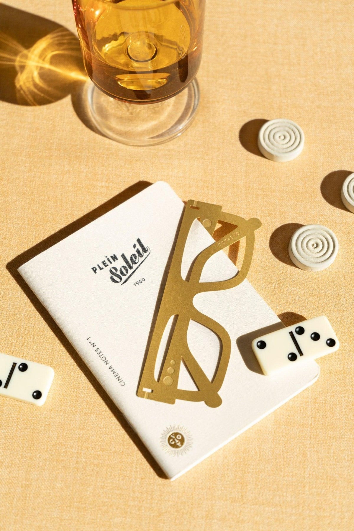 A glass of amber liquid, a book, OCTAEVO gold Bookmark Plein Soleil, dominoes, and game pieces on beige surface.
