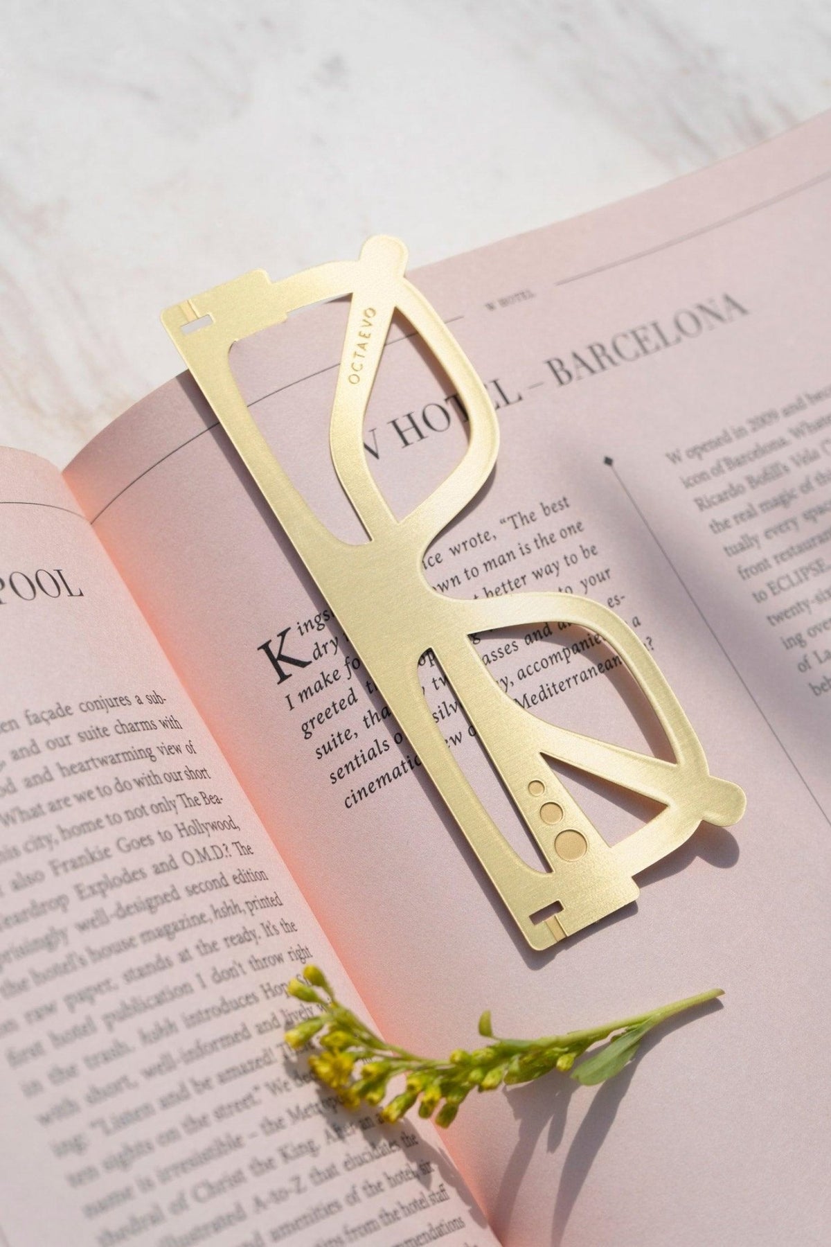 The Plein Soleil Bookmark by OCTAEVO, shaped like gold glasses, lies on a pink book beside a yellow flower.